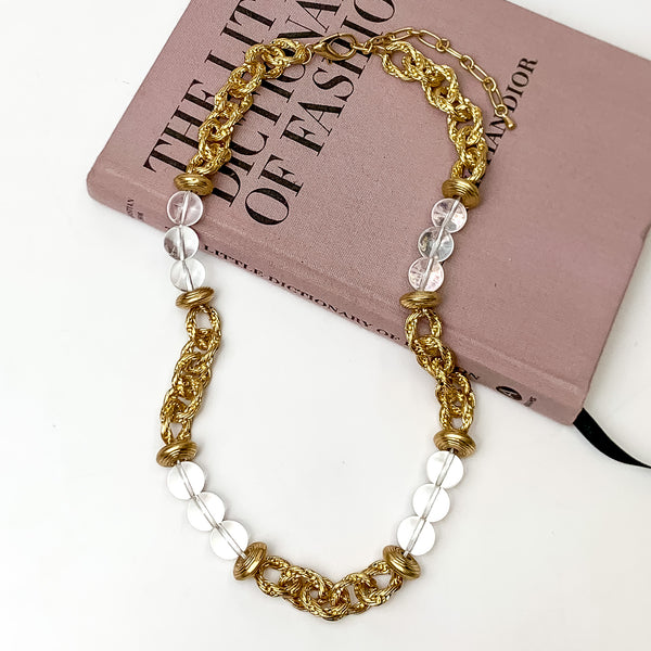 Pictured is a clear beaded and gold chain necklace. This necklace is pictured partially on laying on a mauve colored book on a white background. 