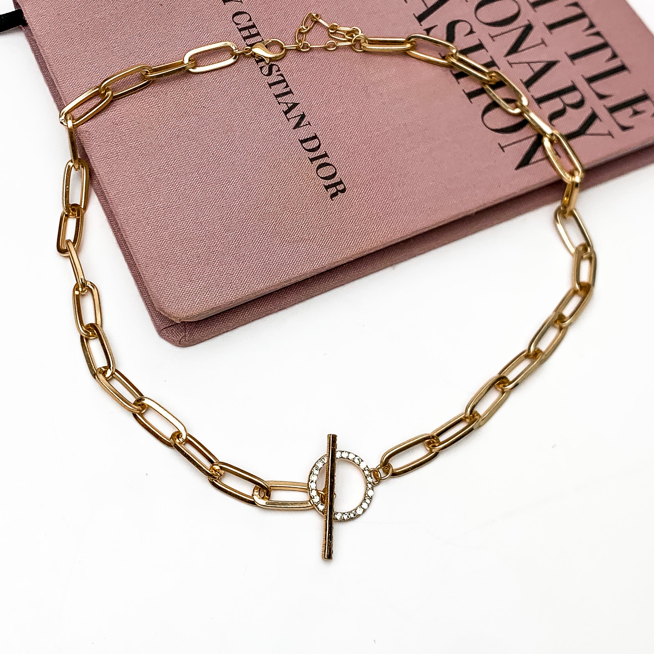 Gold Tone Chain Necklace With Front Toggle. This front toggle gold necklace is on a white background with a pink book behind the necklace.