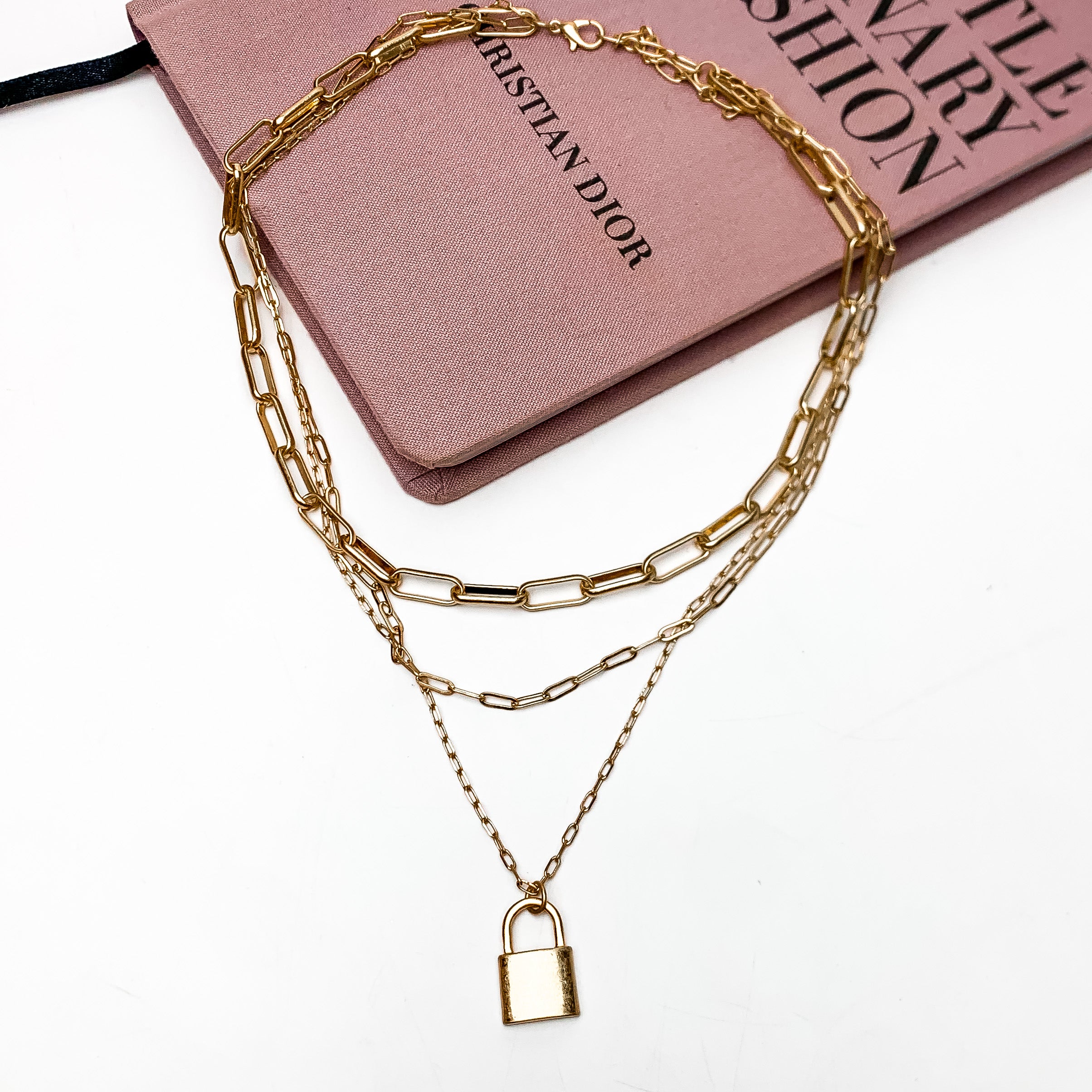 Locked Heart Gold Tone Triple Stand Chain Necklace. This necklace is pictured laying on a pink book with a white background.