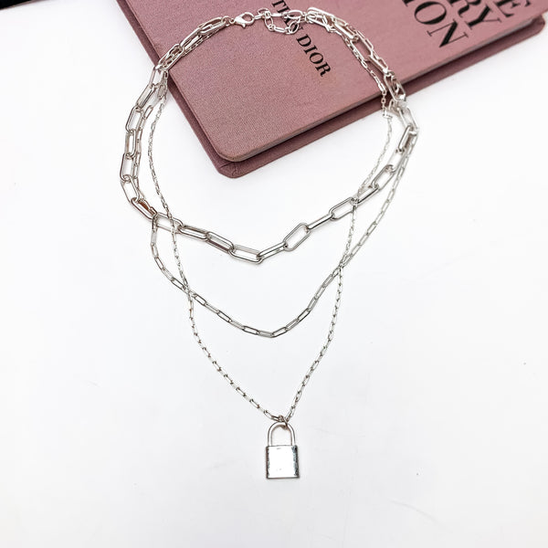 Locked Heart Silver Tone Triple Stand Chain Necklace. This necklace is pictured laying on a pink book with a white background.