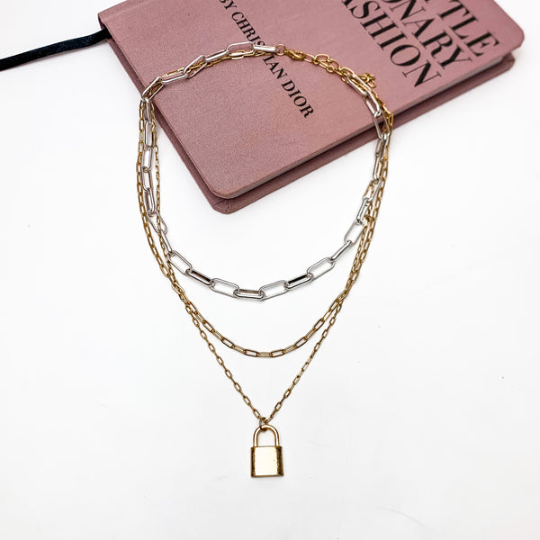 Locked Heart Mixed Metals Triple Stand Chain Necklace. This necklace is pictured laying on a pink book with a white background.