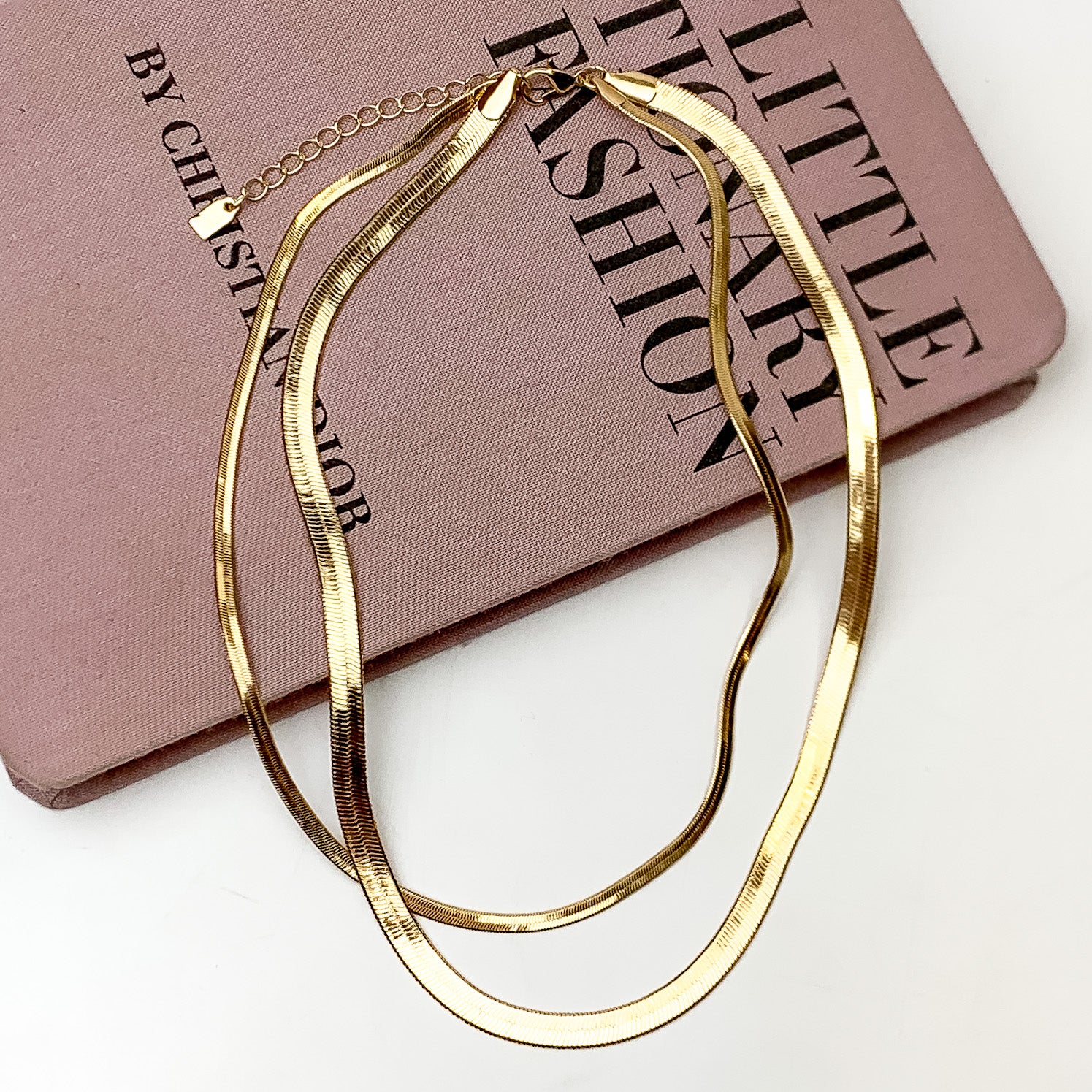 Pictured is a gold, two strand, herringbone chain necklace. This necklace is pictured laying partially on a mauve book on a white background.  