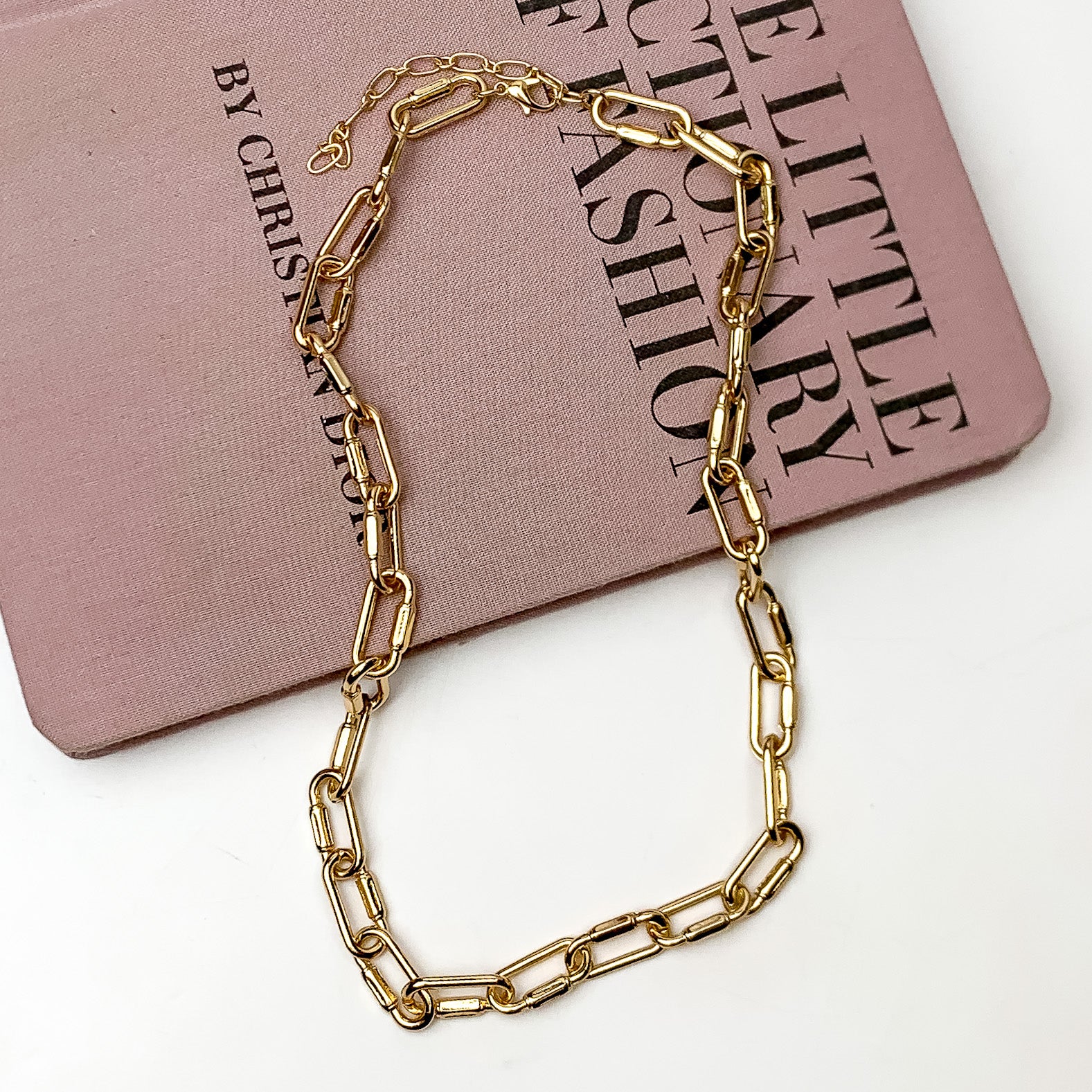 Pictured is a gold chain necklace. This necklace is pictured laying partially on a mauve book on a white background.  