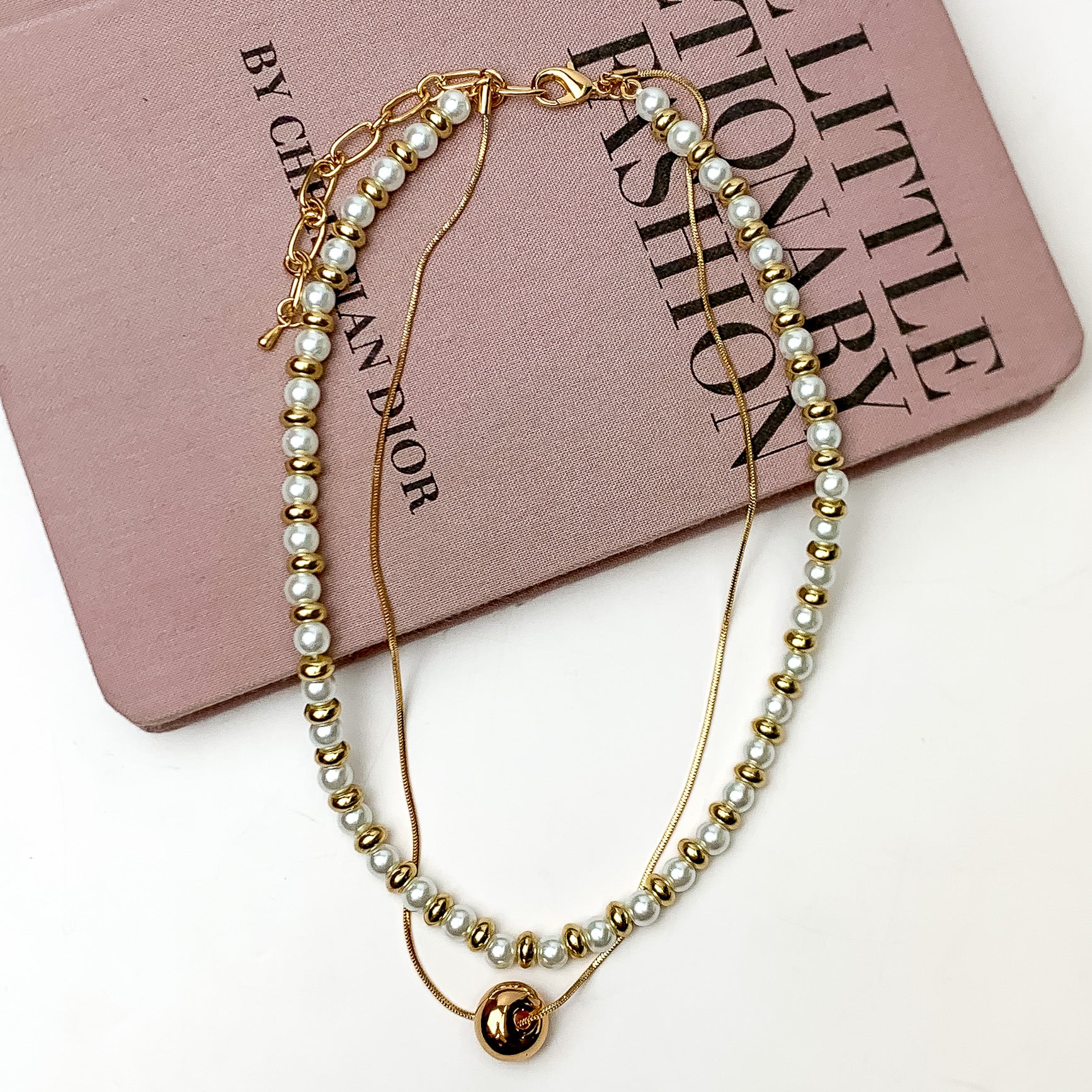 Pictured is a double strand gold and white beaded necklace with a thin strand that has a single gold bead. This necklace is pictured laying partially on a mauve book on a white background.  