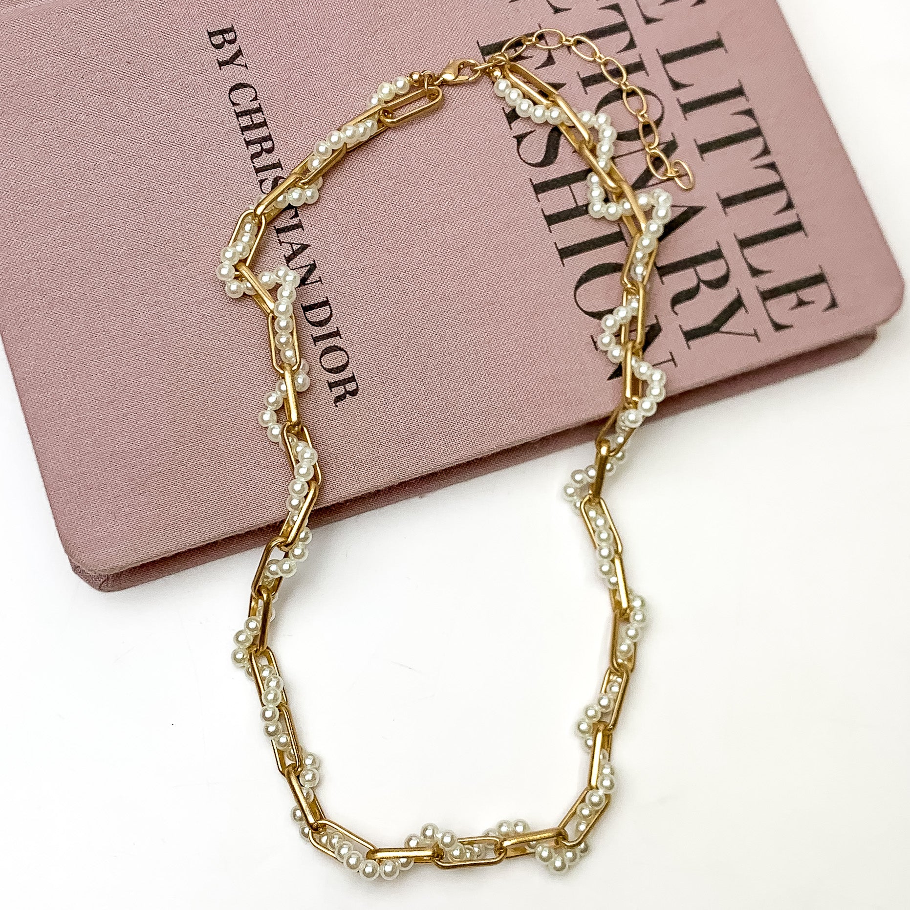 Pictured is a gold chain necklace with a white beaded strand woven through the chain. This necklace is pictured laying partially on a mauve book on a white background.  