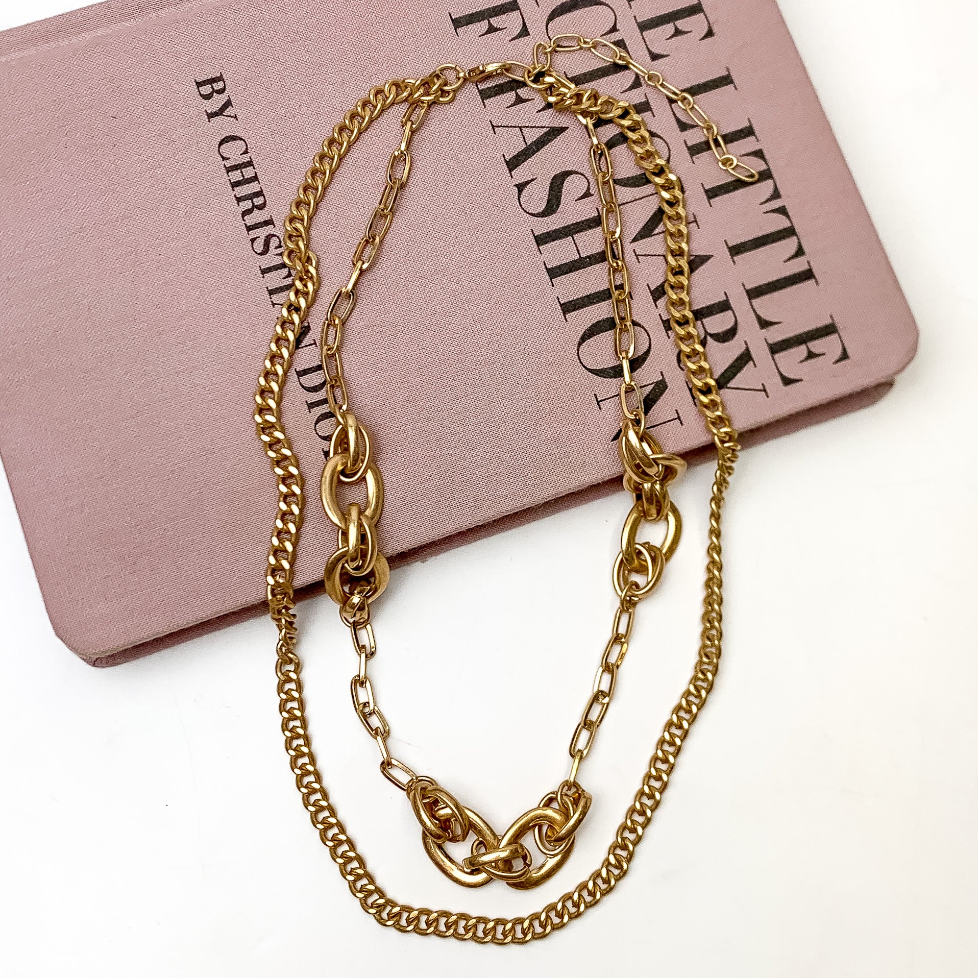 Pictured is a gold, double layered chain necklace. This necklace is pictured laying partially on a mauve book on a white background.  
