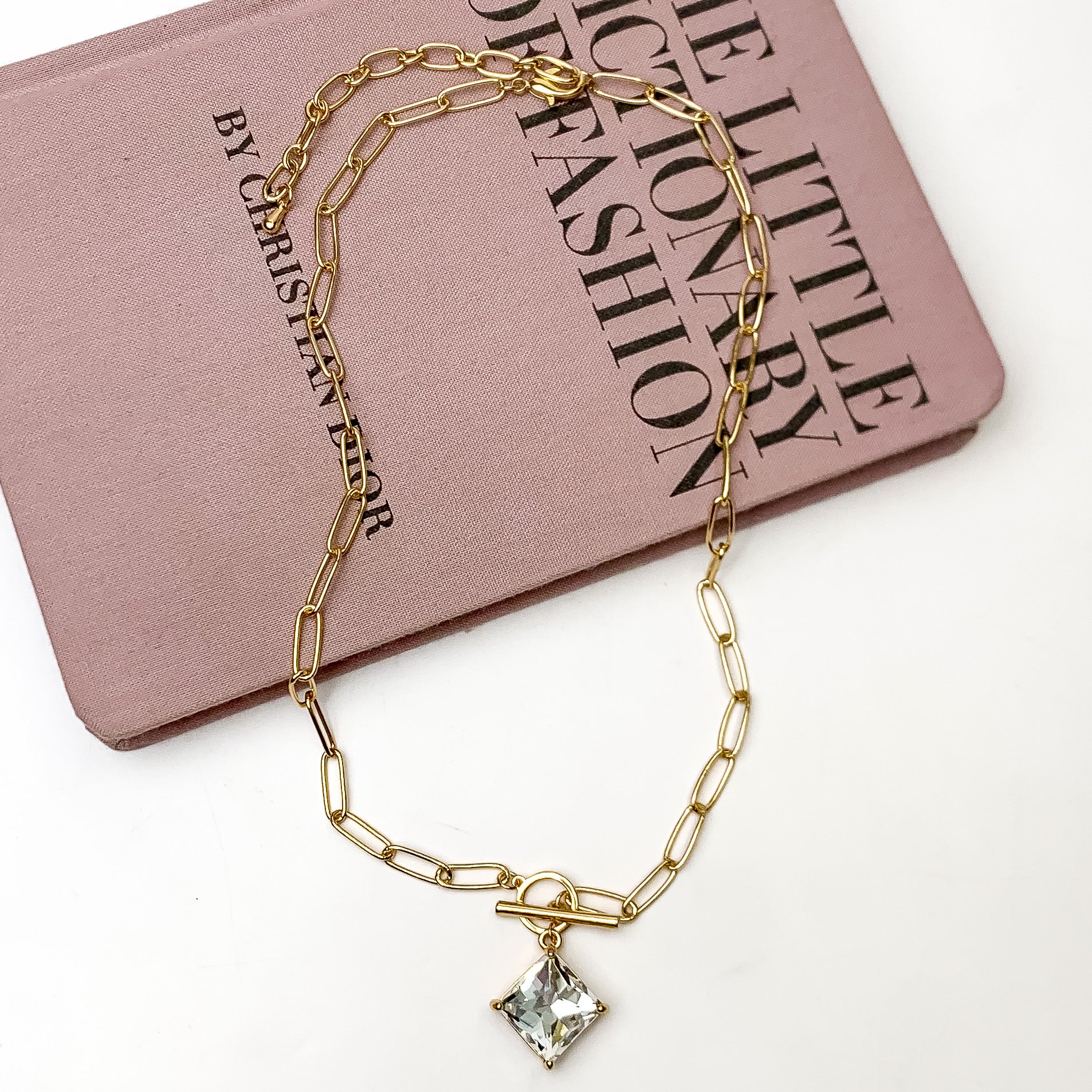 Pictured is a gold, chain necklace with a front toggle clasp and clear, square crystal charm. This necklace is pictured laying partially on a mauve book on a white background.  