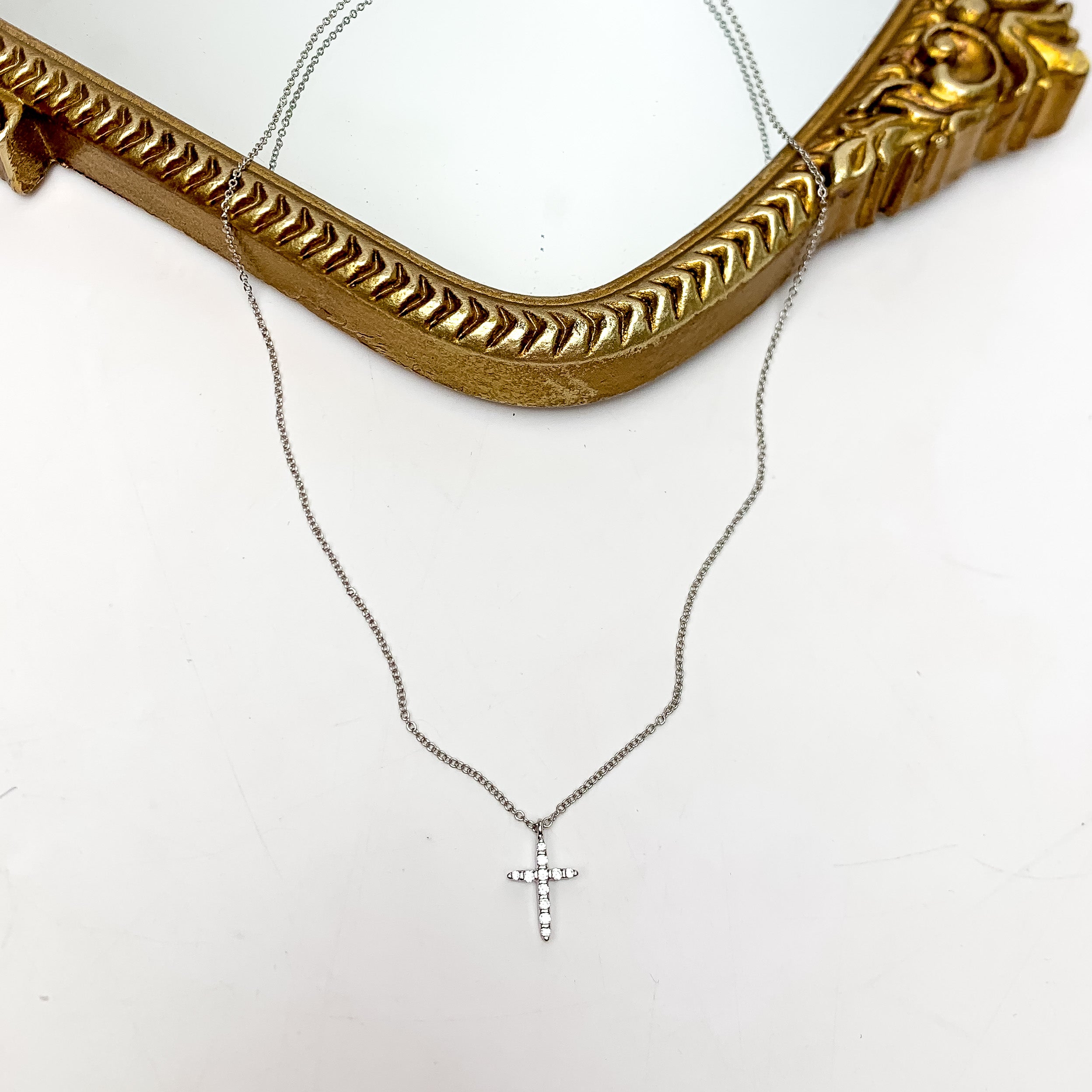 Silver Tone Glory Chain Necklace With Clear Crystal Cross. This necklace is pictured on a white background with part of it on a gold trimmed mirror.