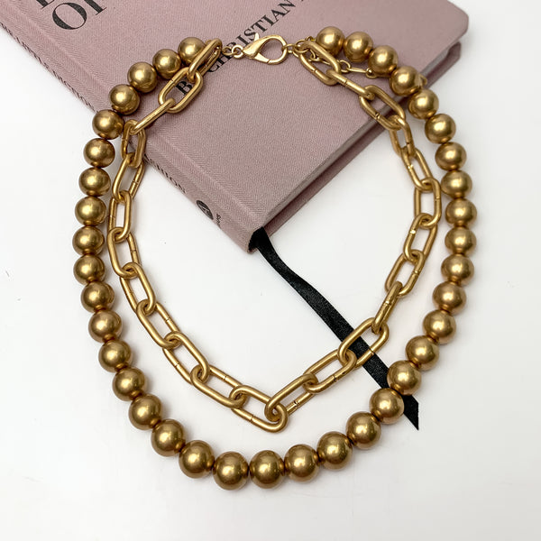Double layered gold tone necklace. One strand is a chain, one strand is beads.. This necklace is pictured on a white background with part of it on a pink book.