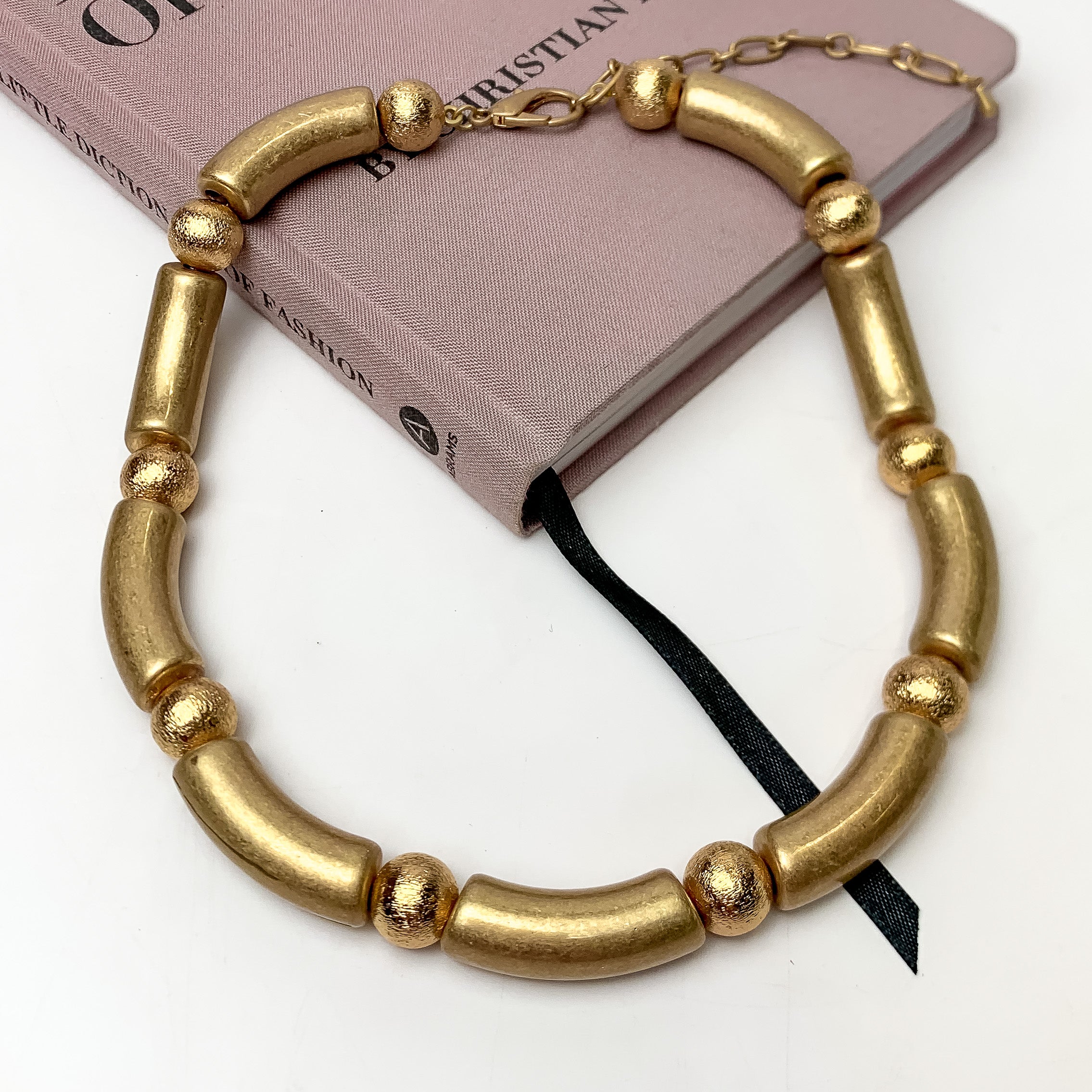 The One To Impress Gold Tone Necklace. This necklace is pictured on a white background with part of it on a pink book.