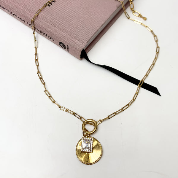 Gold tone chain necklace with circle pendant and clear crystal charm. This necklace is pictured on a white background with part of it on a pink book.