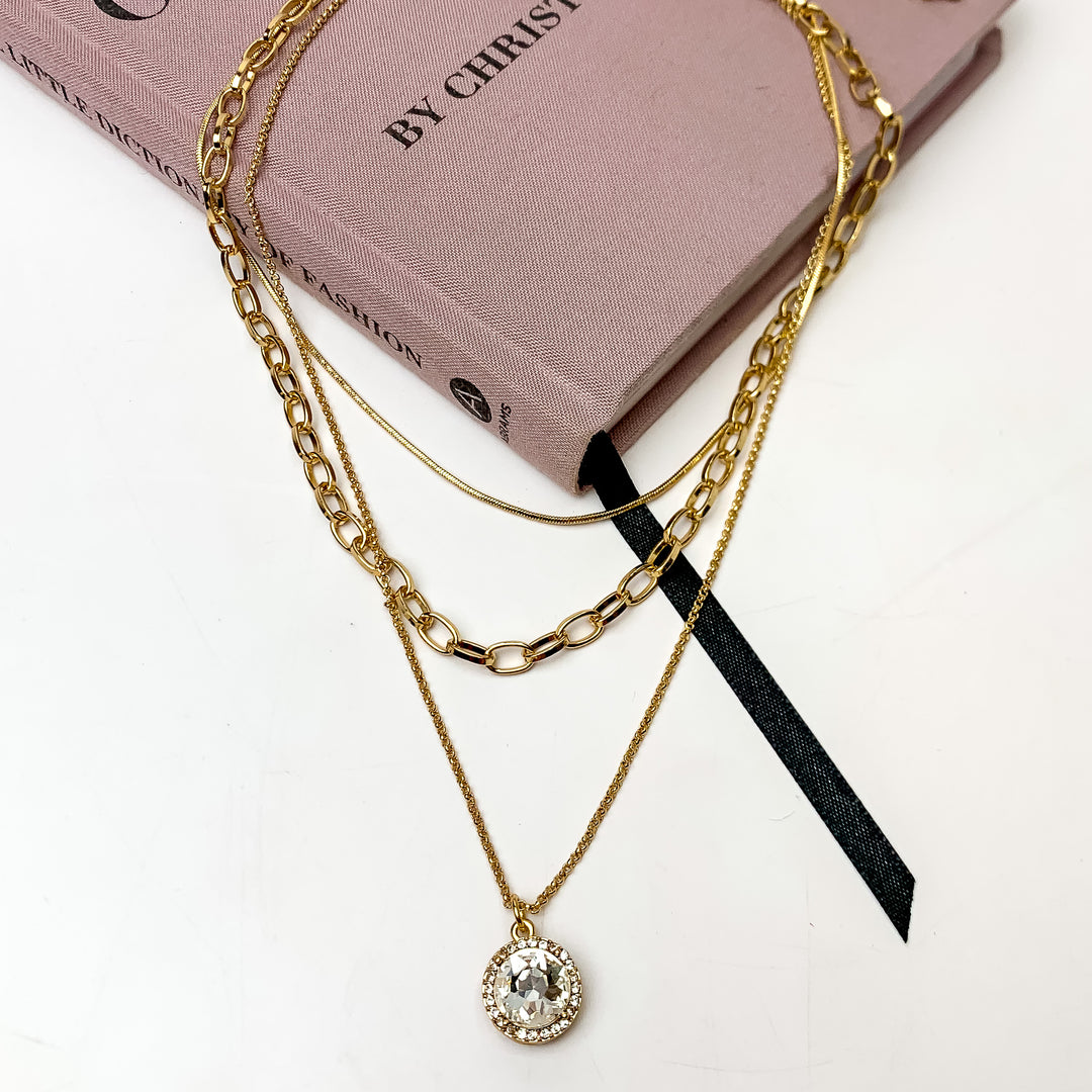 Ready to shine Gold Tone Chain Necklace With Clear Crystal Charm. This necklace is pictured on a white background with part of it on a pink book.