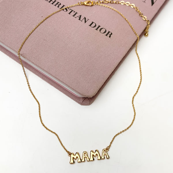 Mama Bubble Letter Chain Necklace in Gold Tone. This necklace is pictured on a white background with part of it on a pink book.