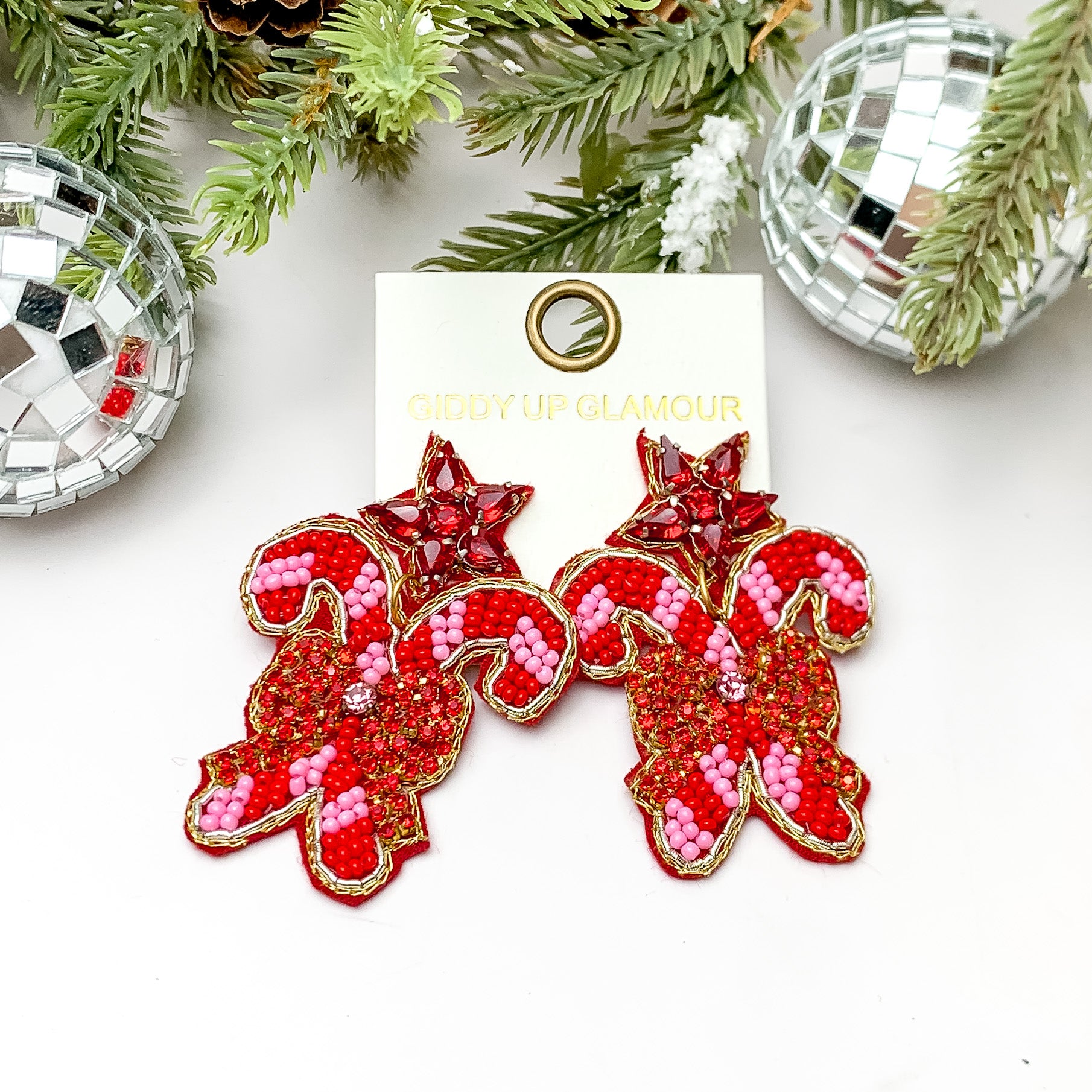 Red and pink candy cane earrings with red crystal bow. These earrings are pictured on a white background with pine trees at the top of the picture. 