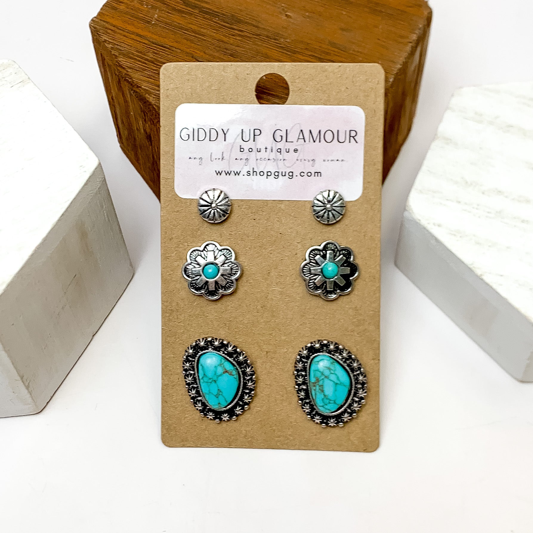 Set Of Three | Flower and Stone Silver Tone Earring Set in Turquoise. These earring are pictured laying against a wooden block with a white background.
