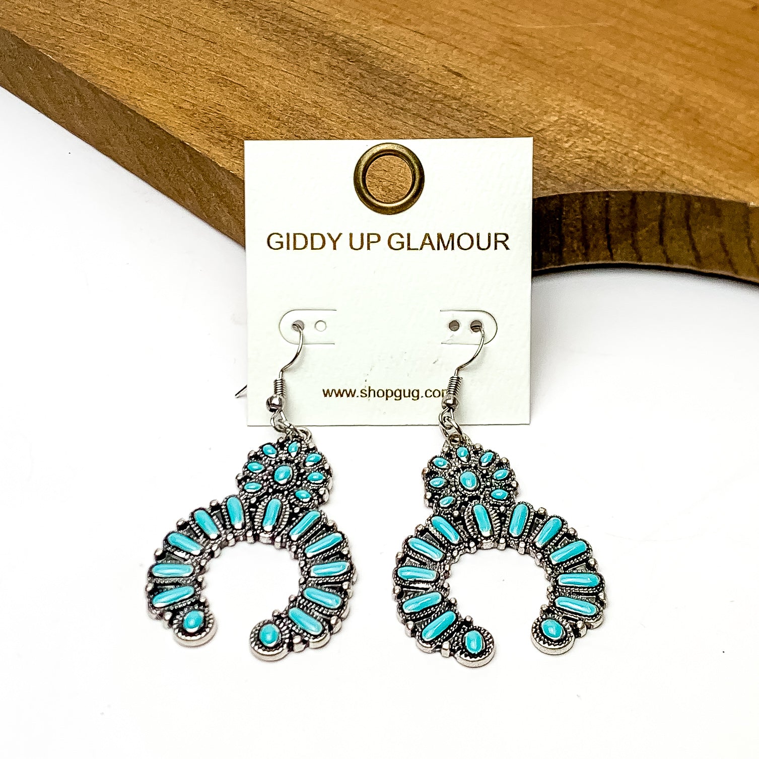 Squash Blossom Silver Tone Medium Earrings in Turquoise. These earrings are pictured on a white background and are laying against a wood piece.
