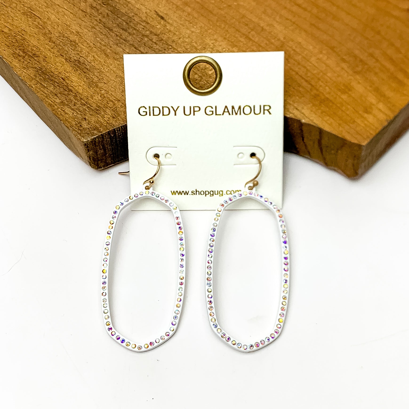 Sparkle Girl Open Oval Earrings in White. These earrings are pictured laying against a wood piece with a white background.