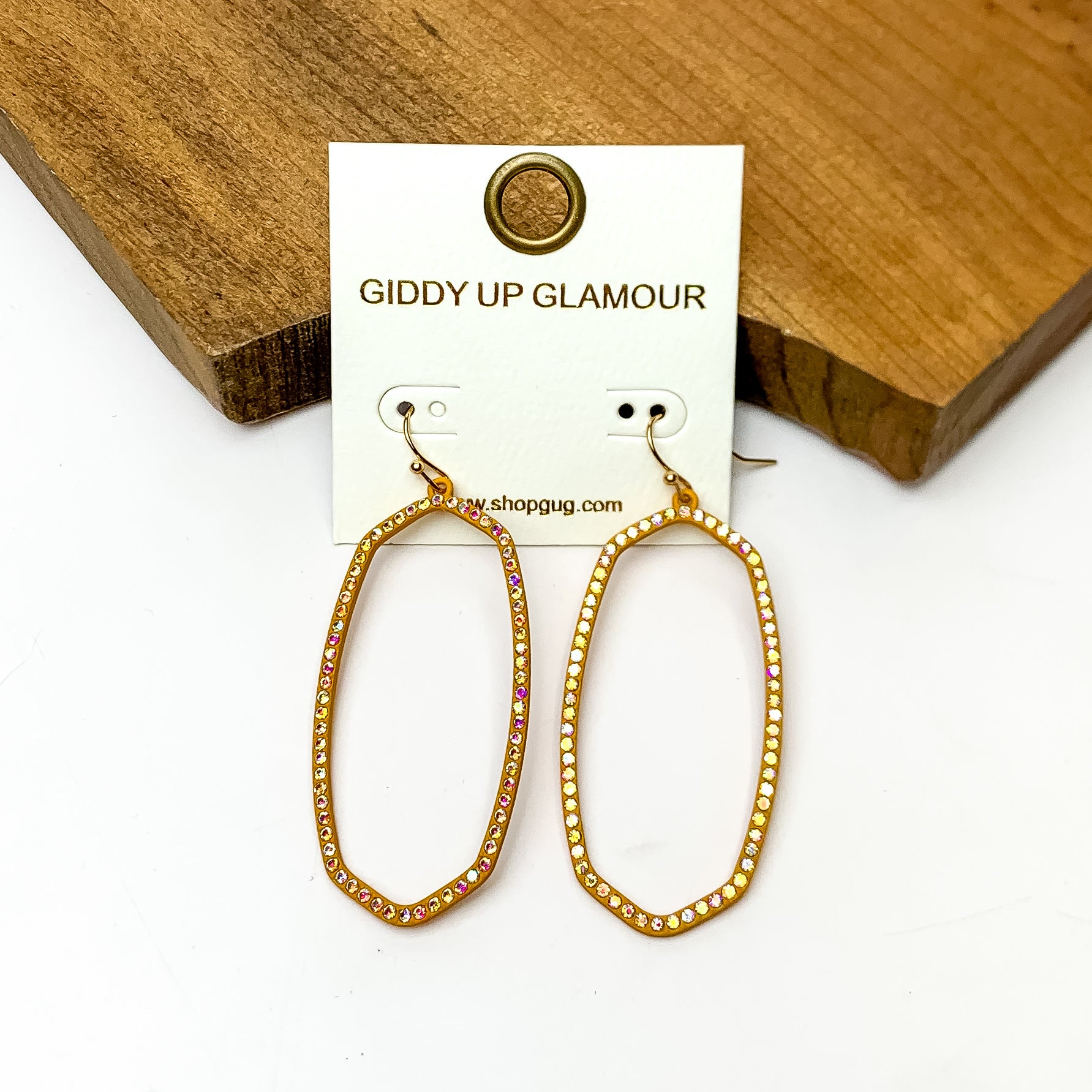 Sparkle Girl Open Oval Earrings in Mustard Yellow. These earrings are pictured laying against a wood piece with a white background.