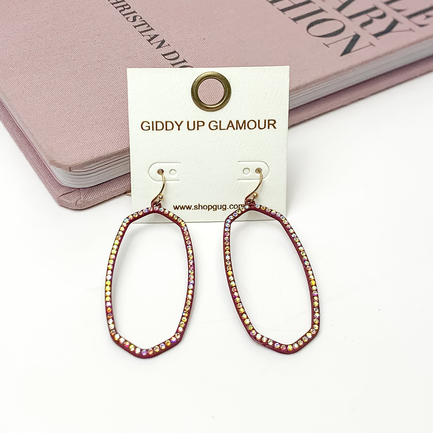Sparkle Girl Open Oval Earrings in Maroon. These earrings are pictured laying against a pink book with a white background.
