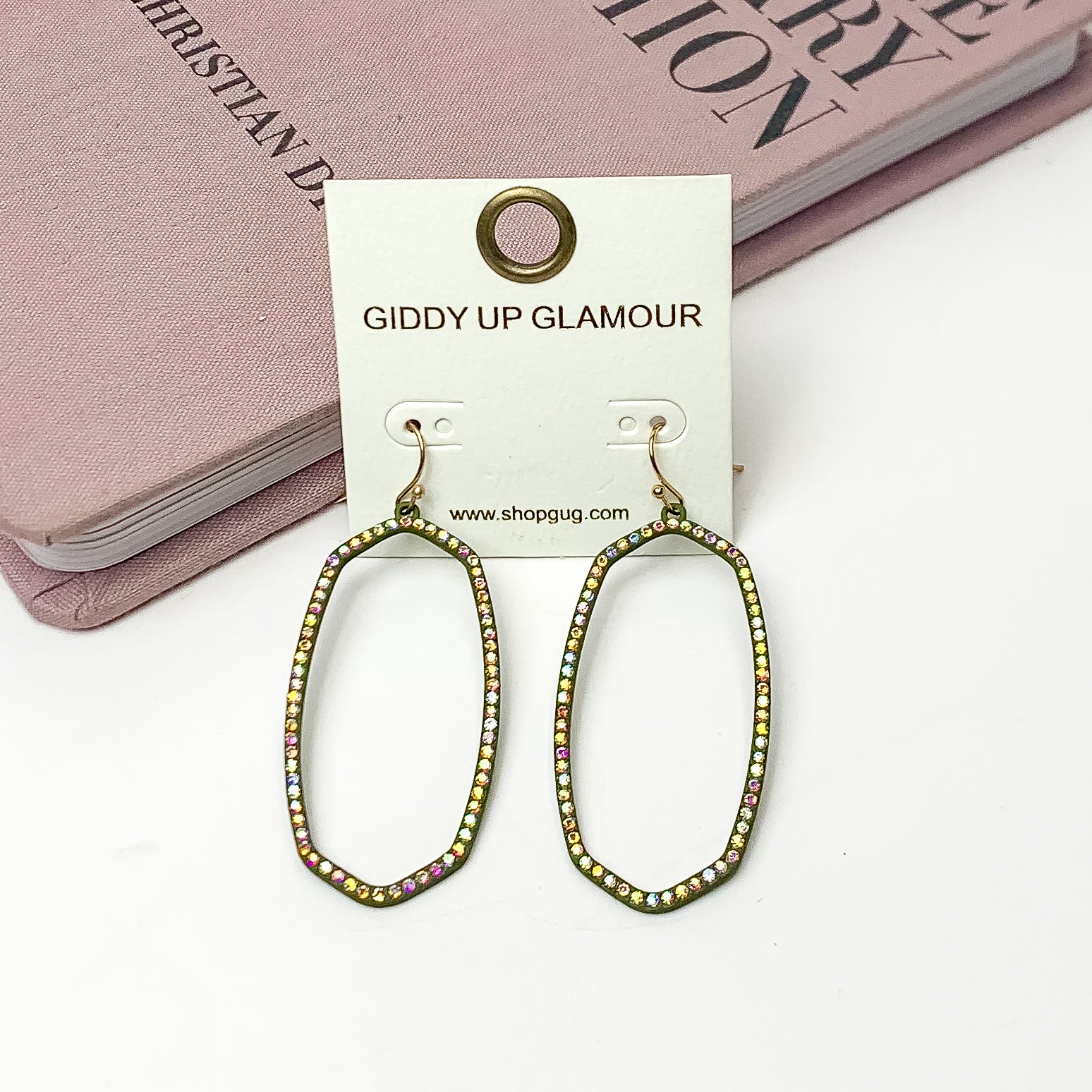 Sparkle Girl Open Oval Earrings in Olive Green. These earrings are pictured laying against a pink book with a white background.