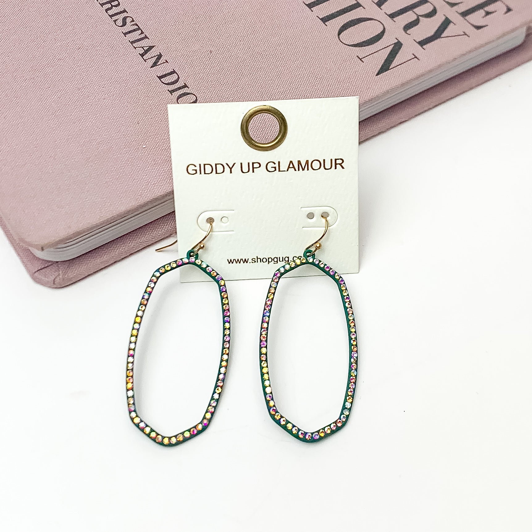 Sparkle Girl Open Oval Earrings in Dark Green. These earrings are pictured laying against a pink book with a white background.