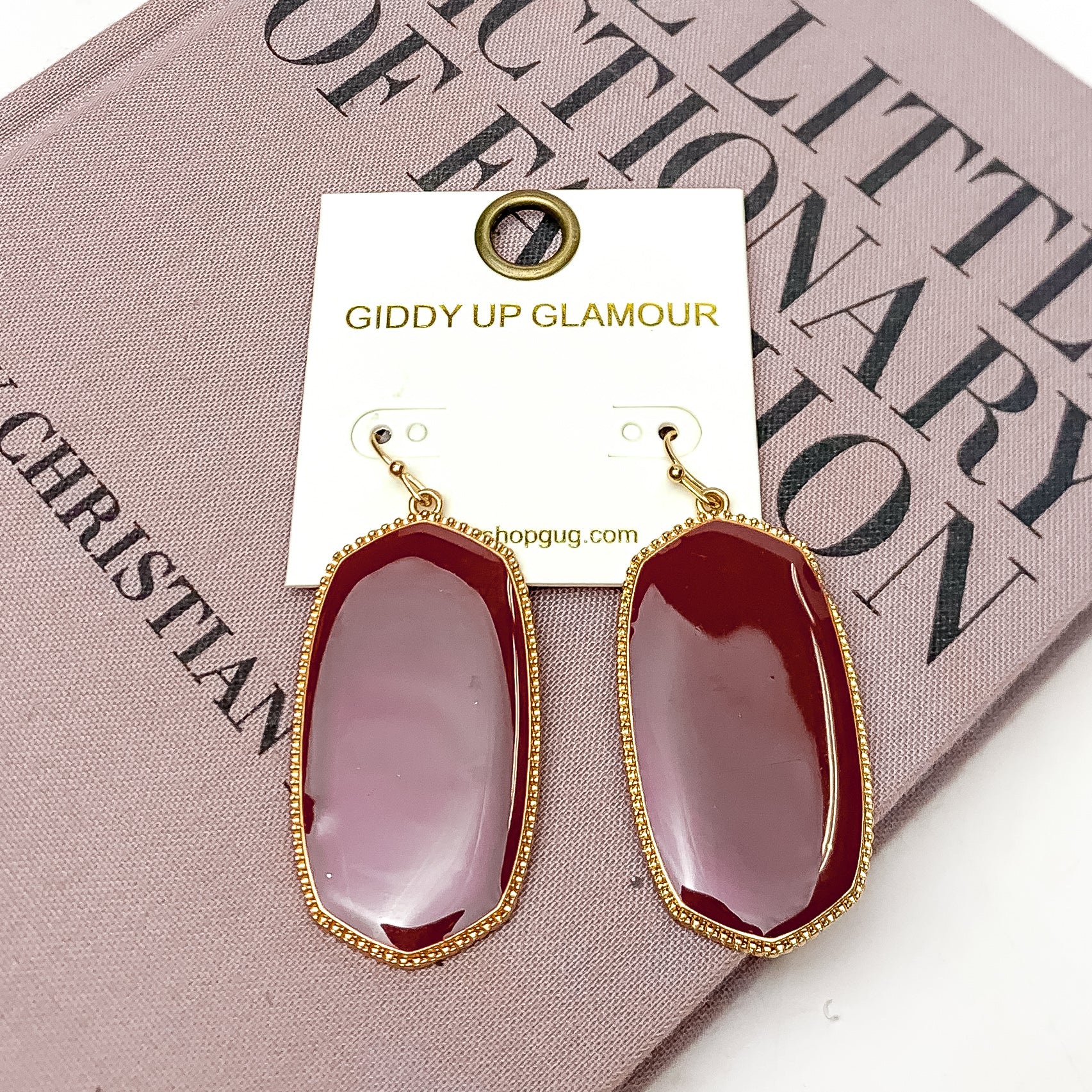 Southern Charm Oval Earrings in Maroon. These earrings are laying on a pink book with a white background behind the book.