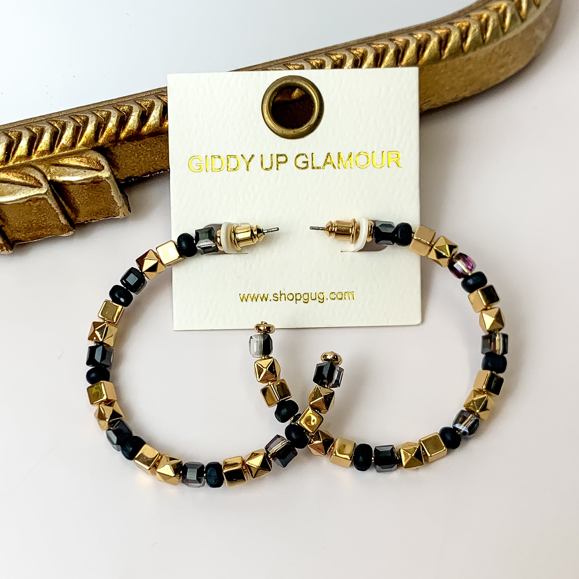 A pair of gold and black beaded hoop earrings on an ivory earring card. These earrings are pictured on a white background with a gold mirror.