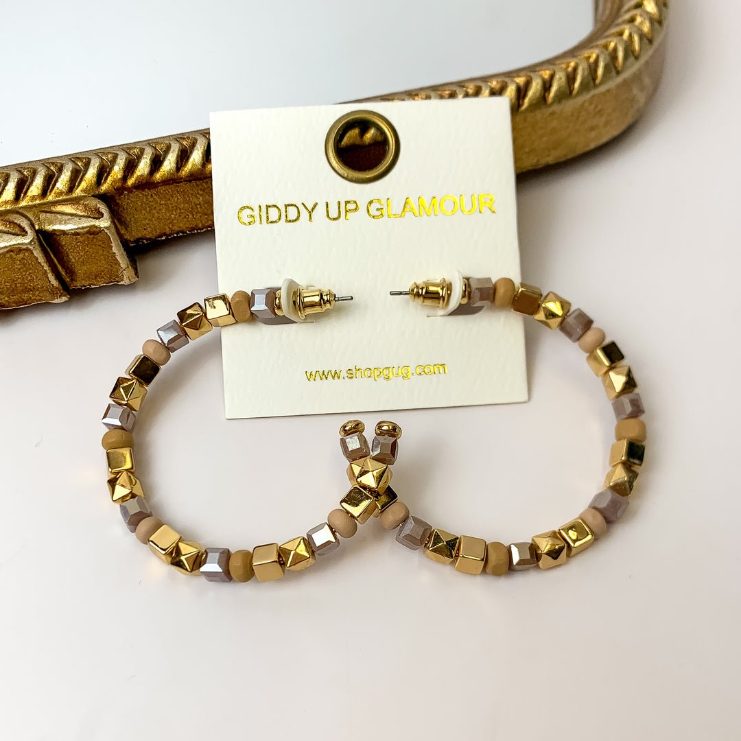 A pair of gold and taupe beaded hoop earrings on an ivory earring card. These earrings are pictured on a white background with a gold mirror.