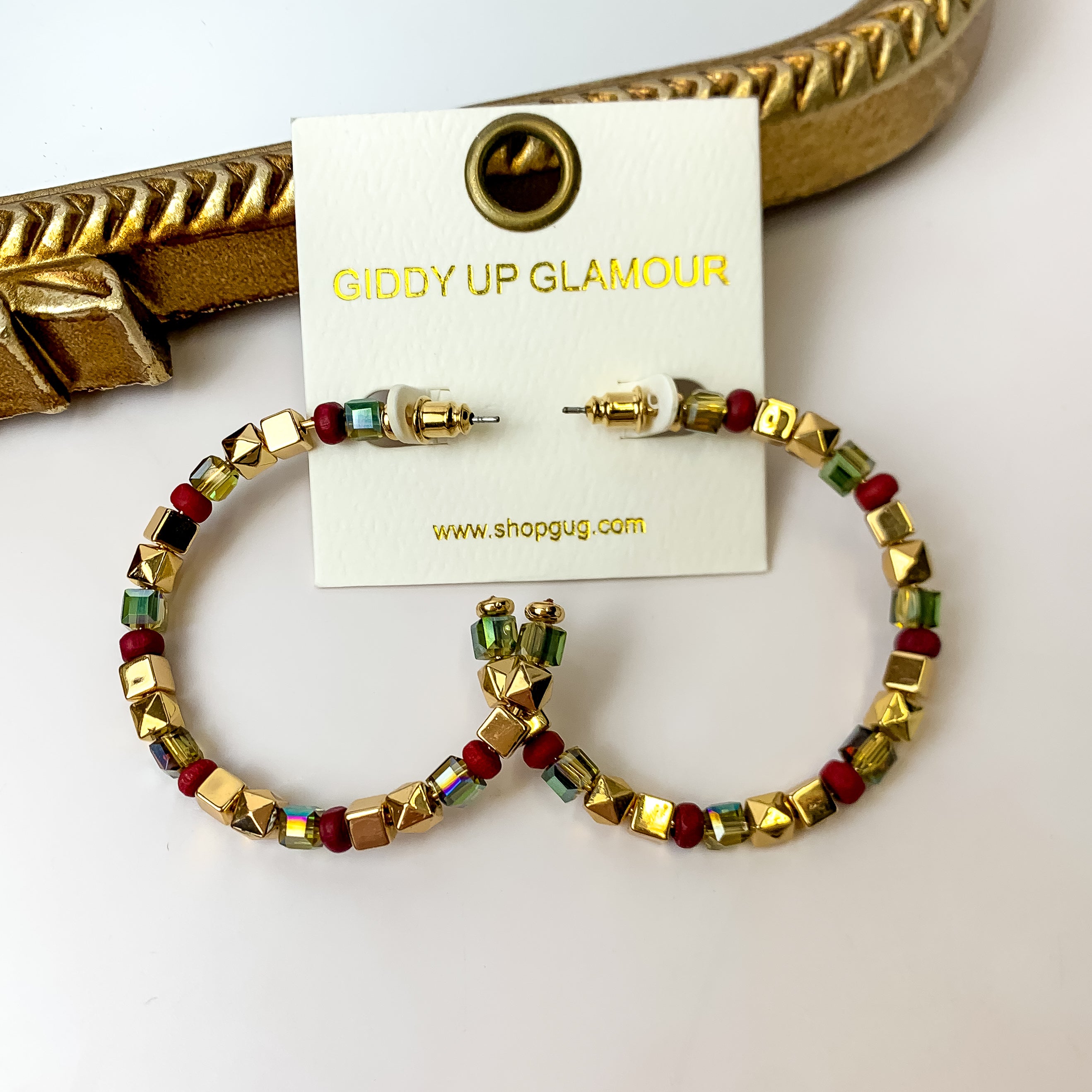 A pair of gold, sage green, and ruby red beaded hoop earrings on an ivory earring card. These earrings are pictured on a white background with a gold mirror.