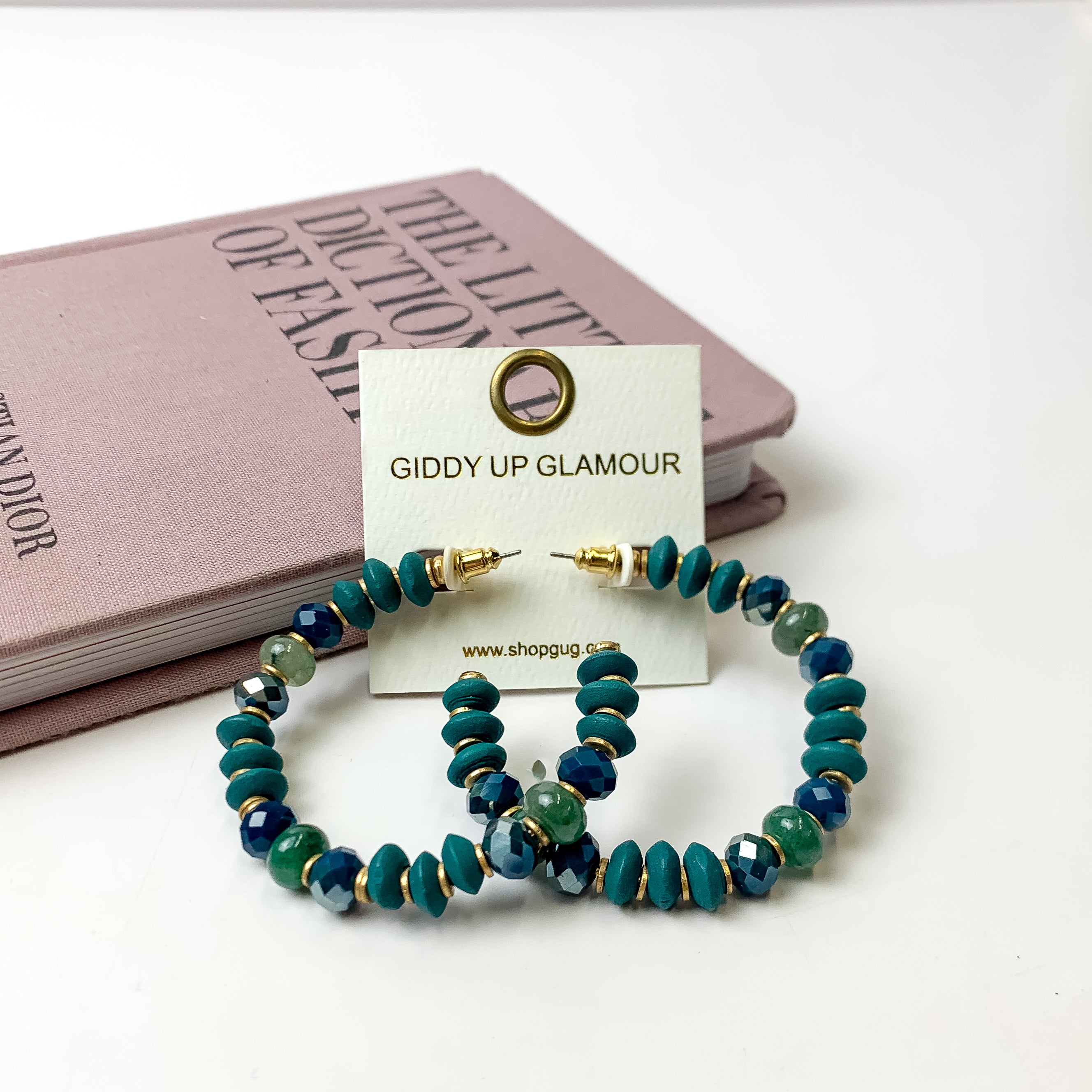 A pair of deep sea blue beaded hoop earrings on an ivory earring card. These earrings are pictured on a white background with  a light purple book. 