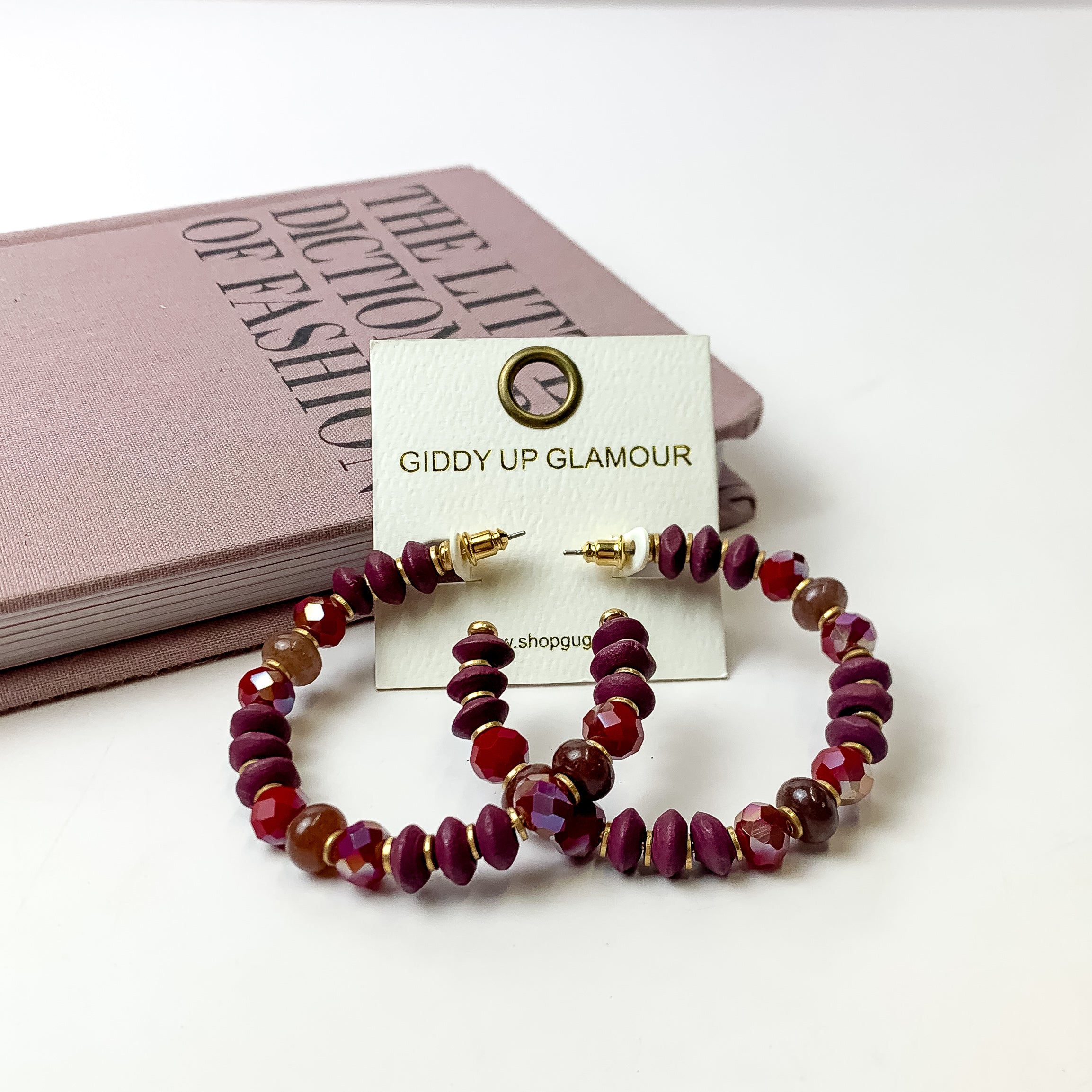 A pair of plum purple beaded hoop earrings on an ivory earring card. These earrings are pictured on a white background with a light purple book!