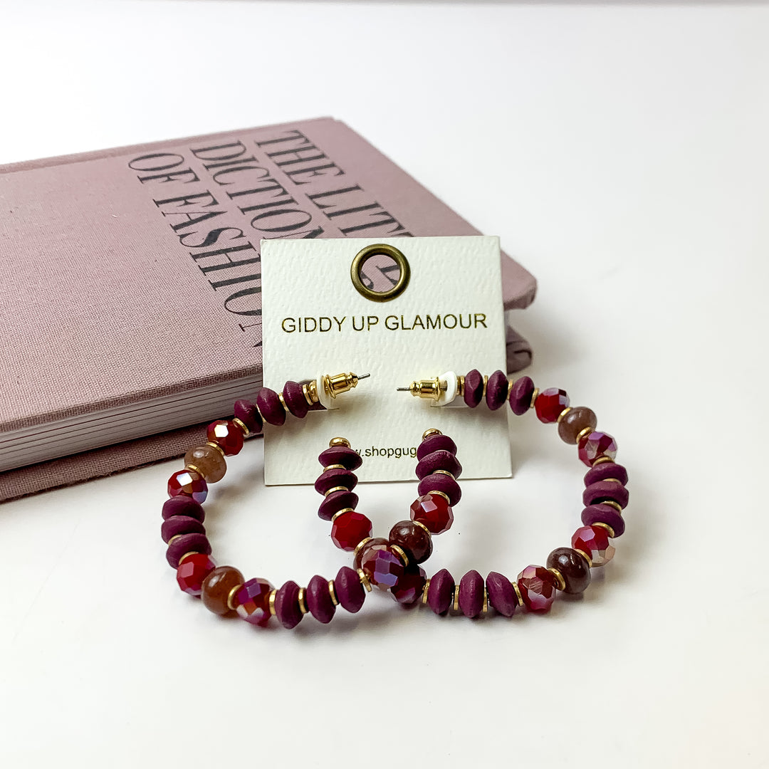 A pair of plum purple beaded hoop earrings on an ivory earring card. These earrings are pictured on a white background with a light purple book!