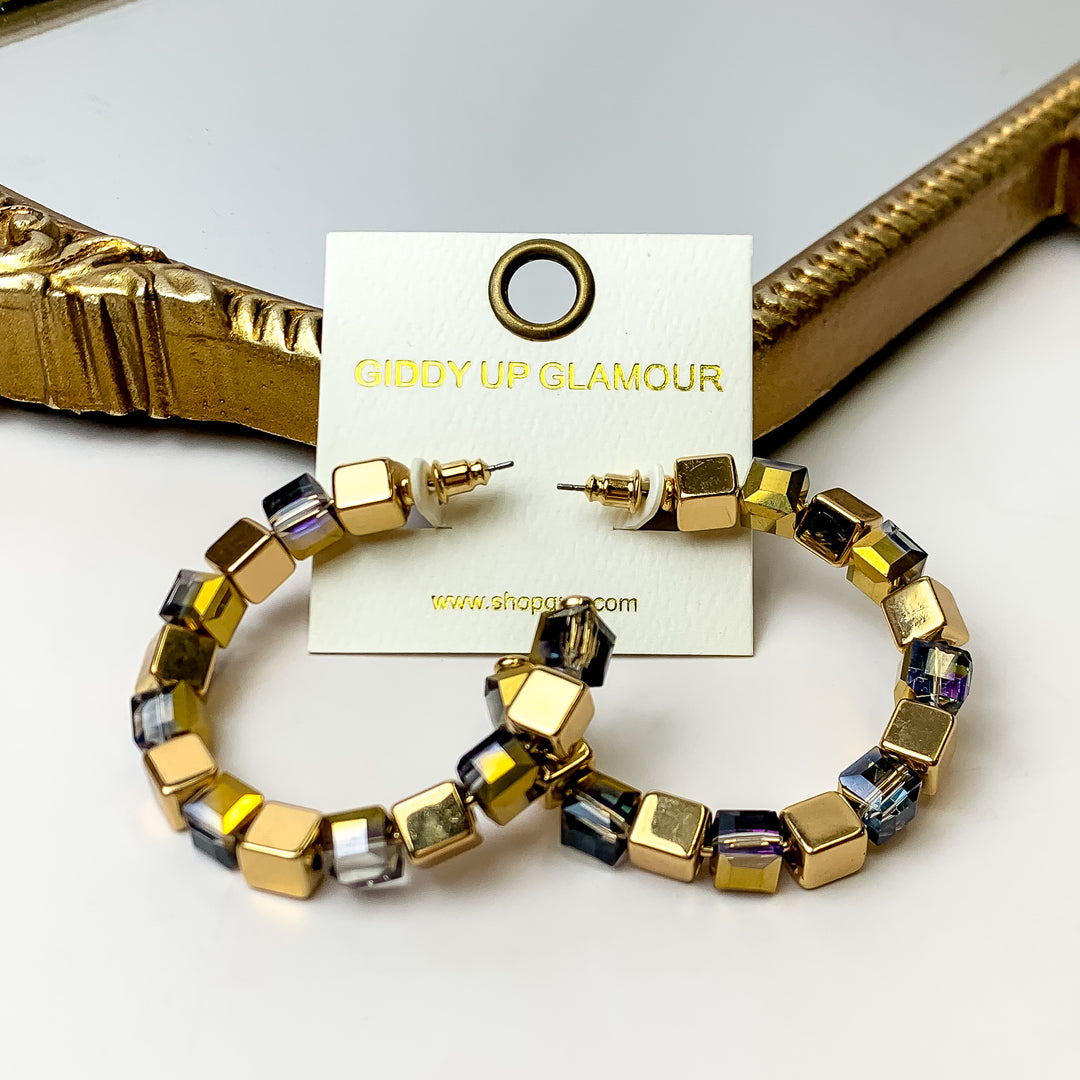 A pair of iridescent purple and gold cubed hoop earrings on an ivory earring card. These earrings are placed against a gold mirror with a white background.