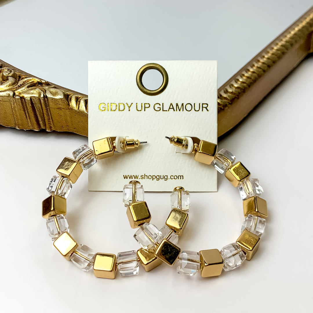 A pair of clear and gold cubed hoop earrings on an ivory earring card. These earrings are placed against a gold mirror with a white background.