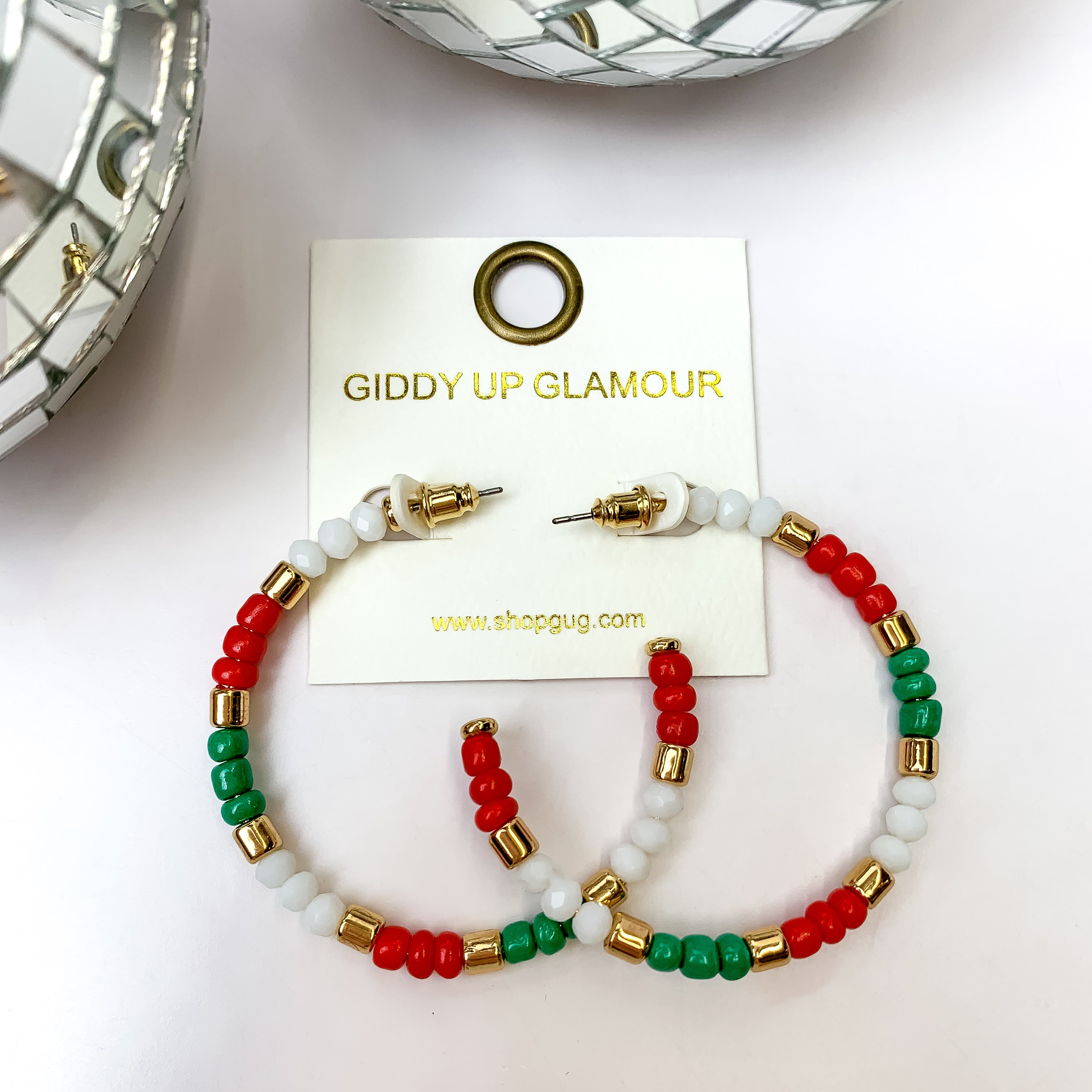 A pair of gold tone, red, white, and green hoop earrings on an ivory earring card. These earrings are pictured with disco balls and a white background.