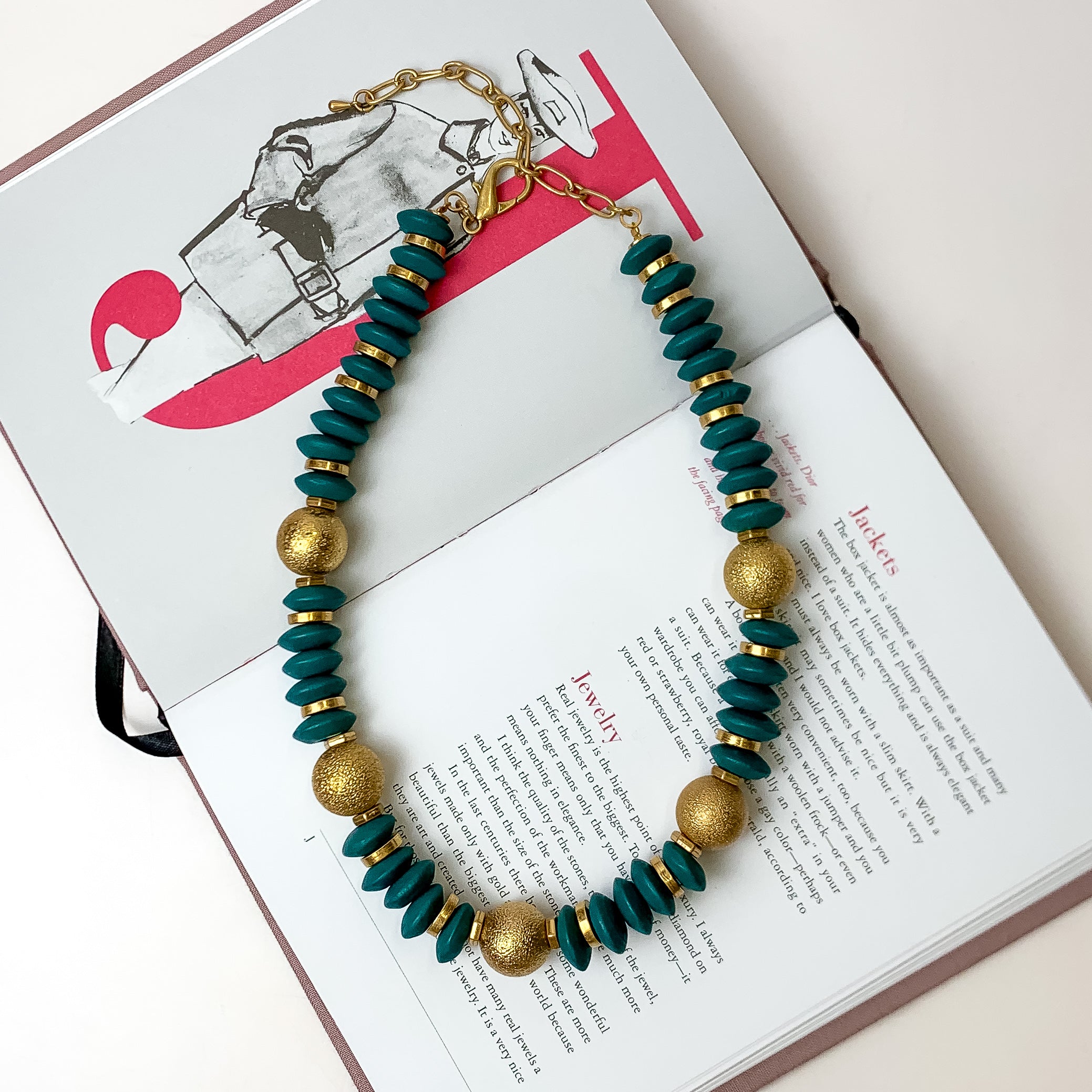 This beaded necklace with gold spacers in the color teal is placed inside the pages of a book with a white background.
