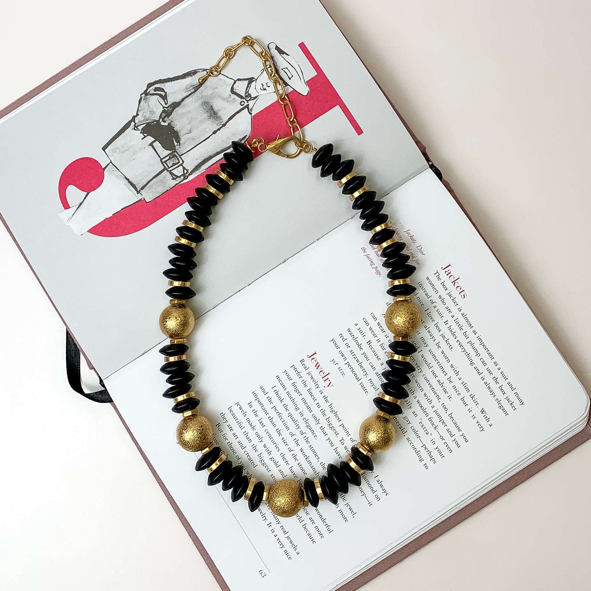 This beaded necklace with gold spacers in the color black is placed inside the pages of a book with a white background.