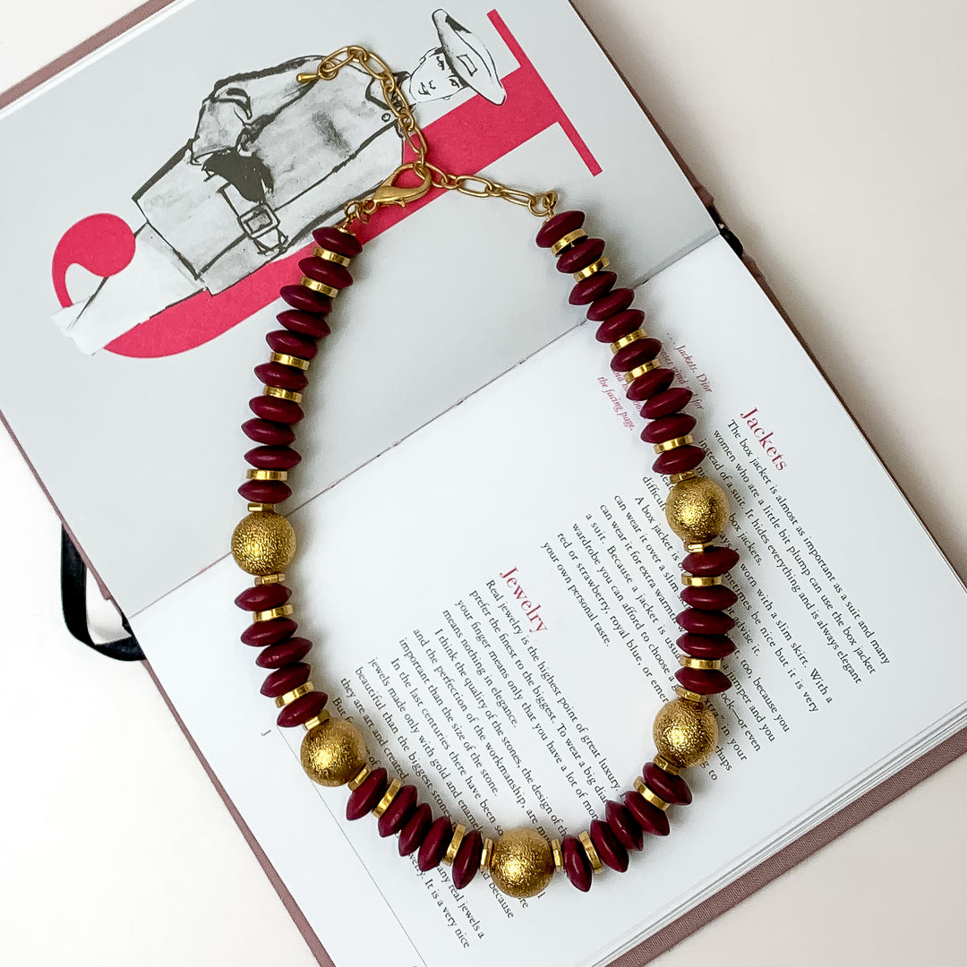 This beaded necklace with gold spacers in the color plum purple is placed inside the pages of a book with a white background.