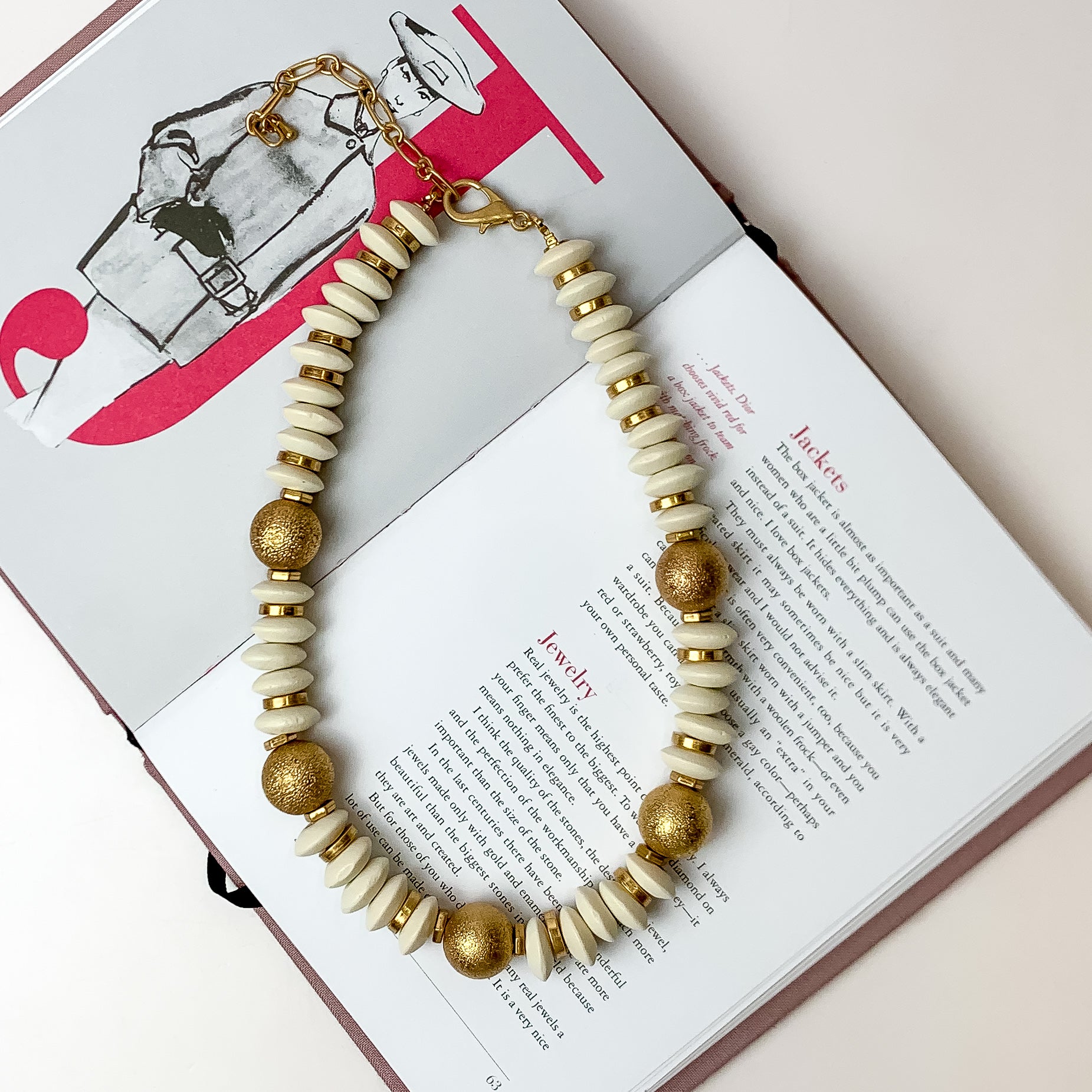 This beaded necklace with gold spacers in the color white is placed inside the pages of a book with a white background.