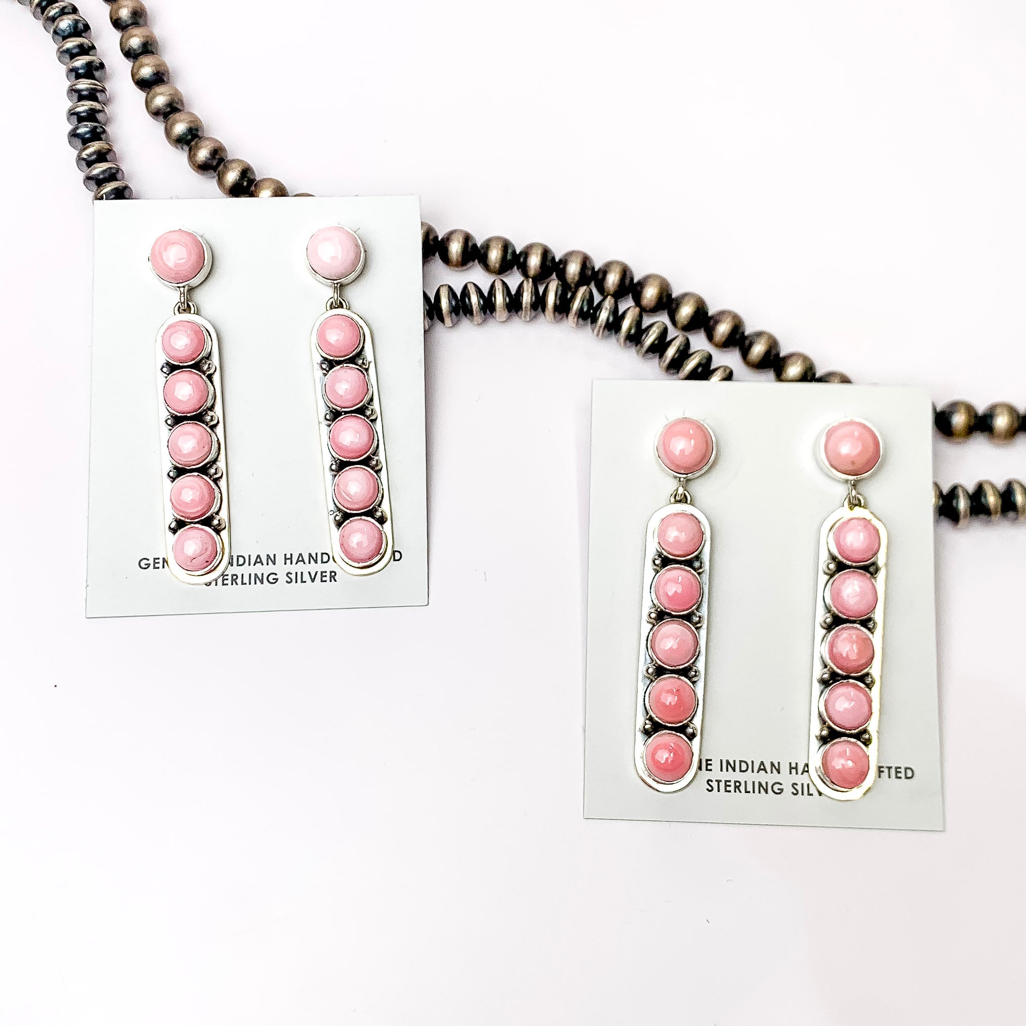Centered in the picture are two sets of pink concho post back earrings. A strand of navajo pearls are laid above the earrings, all on a white background. 