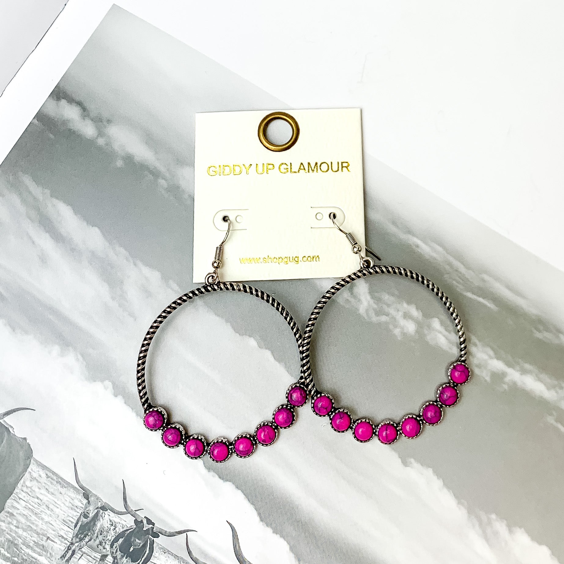 Forever Twisted Hoop Earrings with Stones in Pink. Pictured on a book with a western scene in the background.