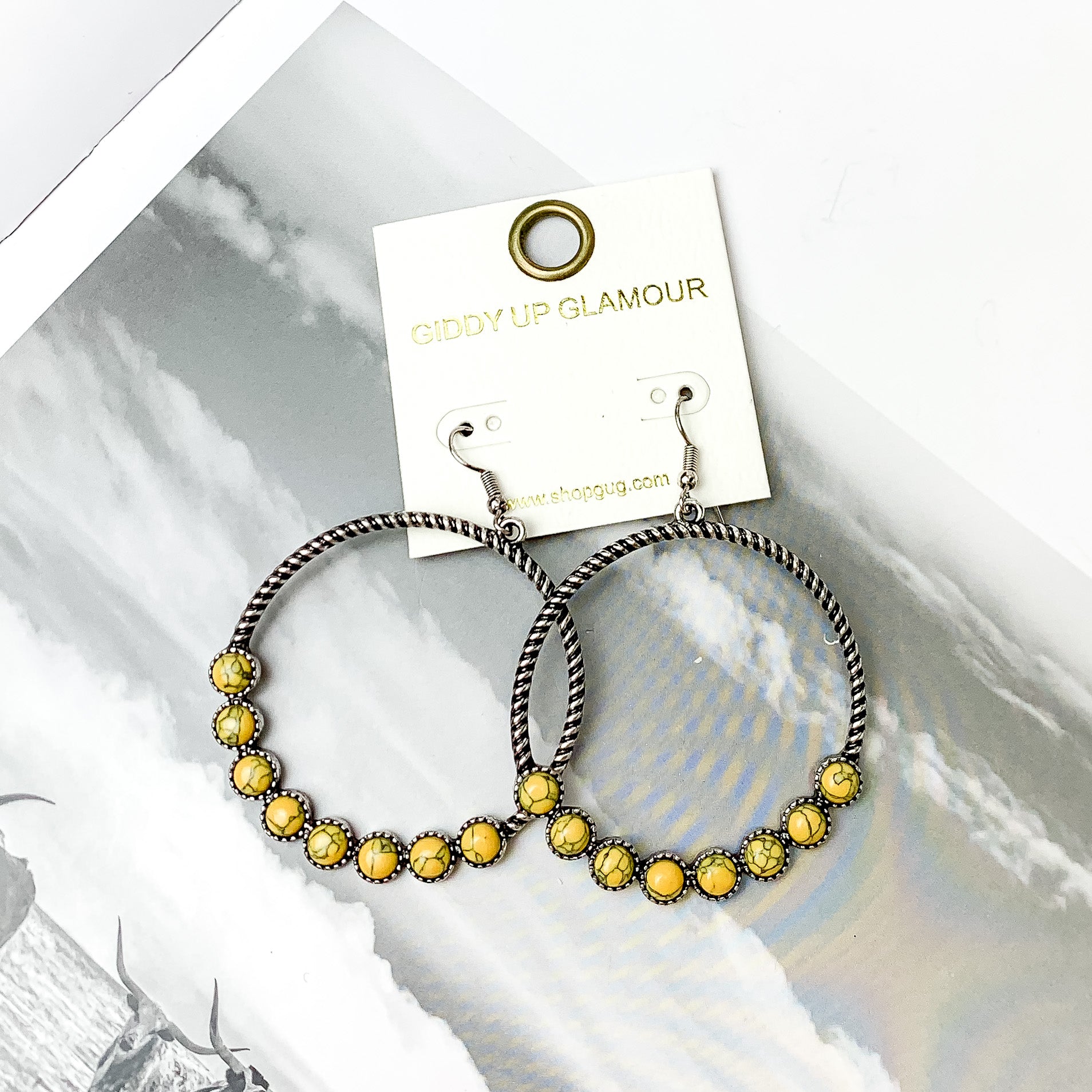 Forever Twisted Hoop Earrings with Stones in Yellow. Pictured on an open book with a gray sky in the background.