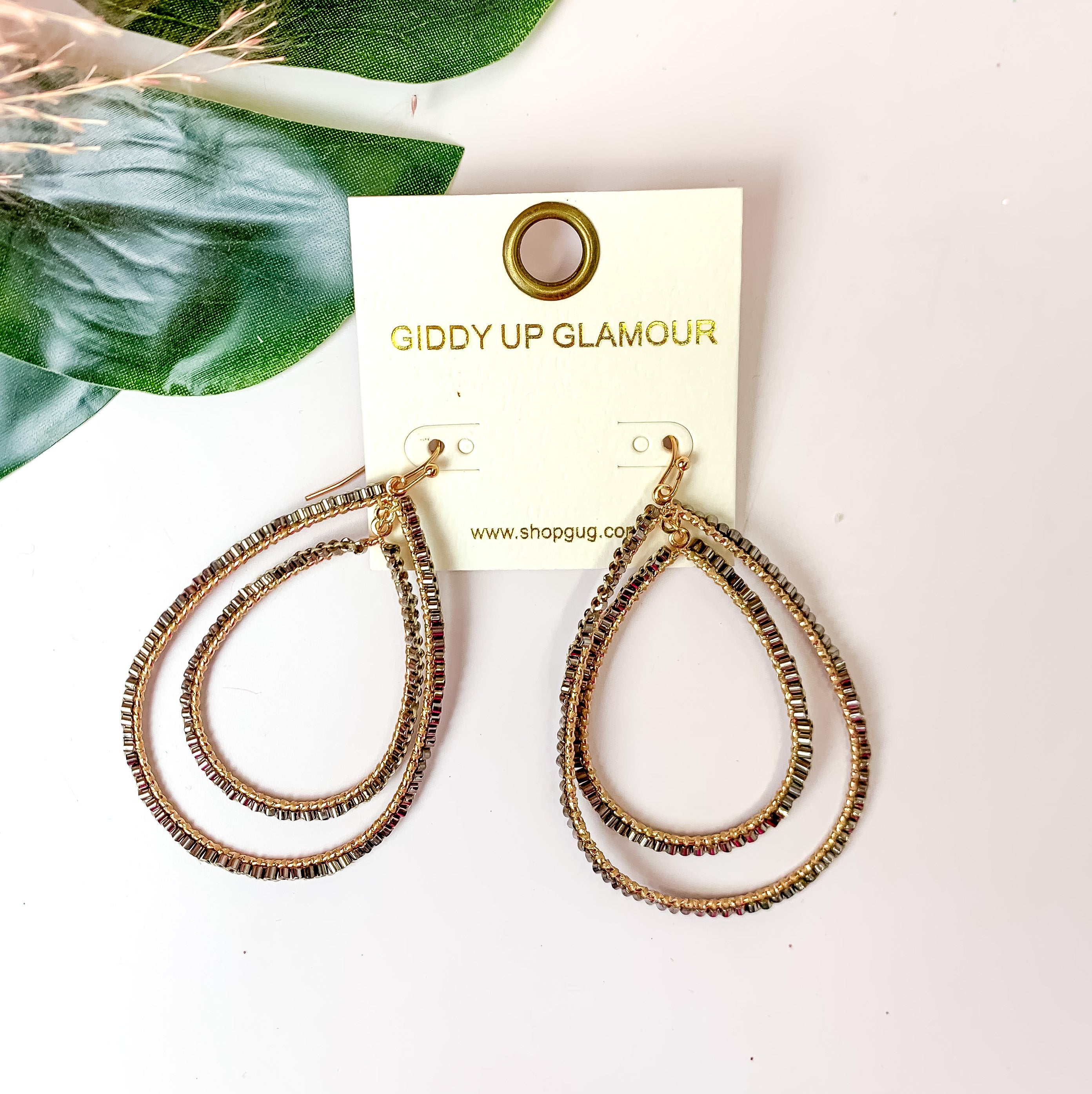 Double Open Teardrop Gold Tone Earrings with Beaded Outline in Charcoal. Pictured on a white background with plants in the top left corner.