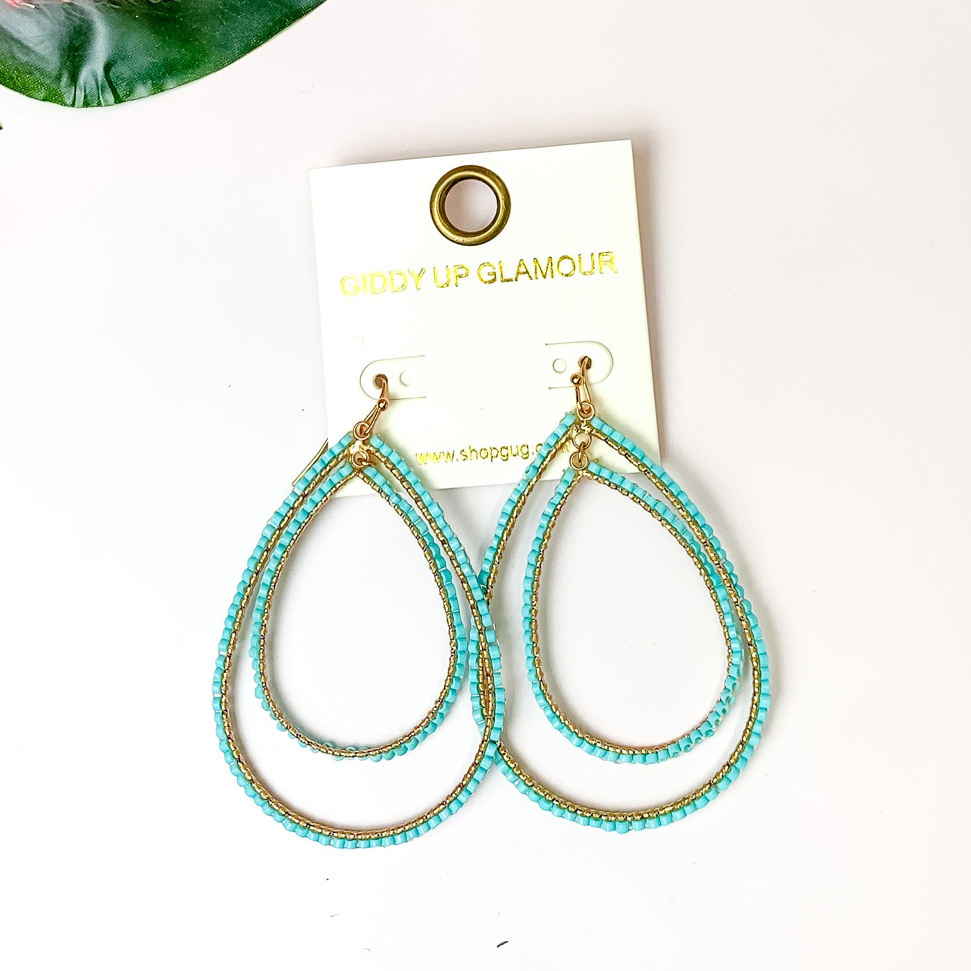Double Open Teardrop Gold Tone Earrings with Beaded Outline in Turquoise. Pictured on a white background with plants in the top left corner.