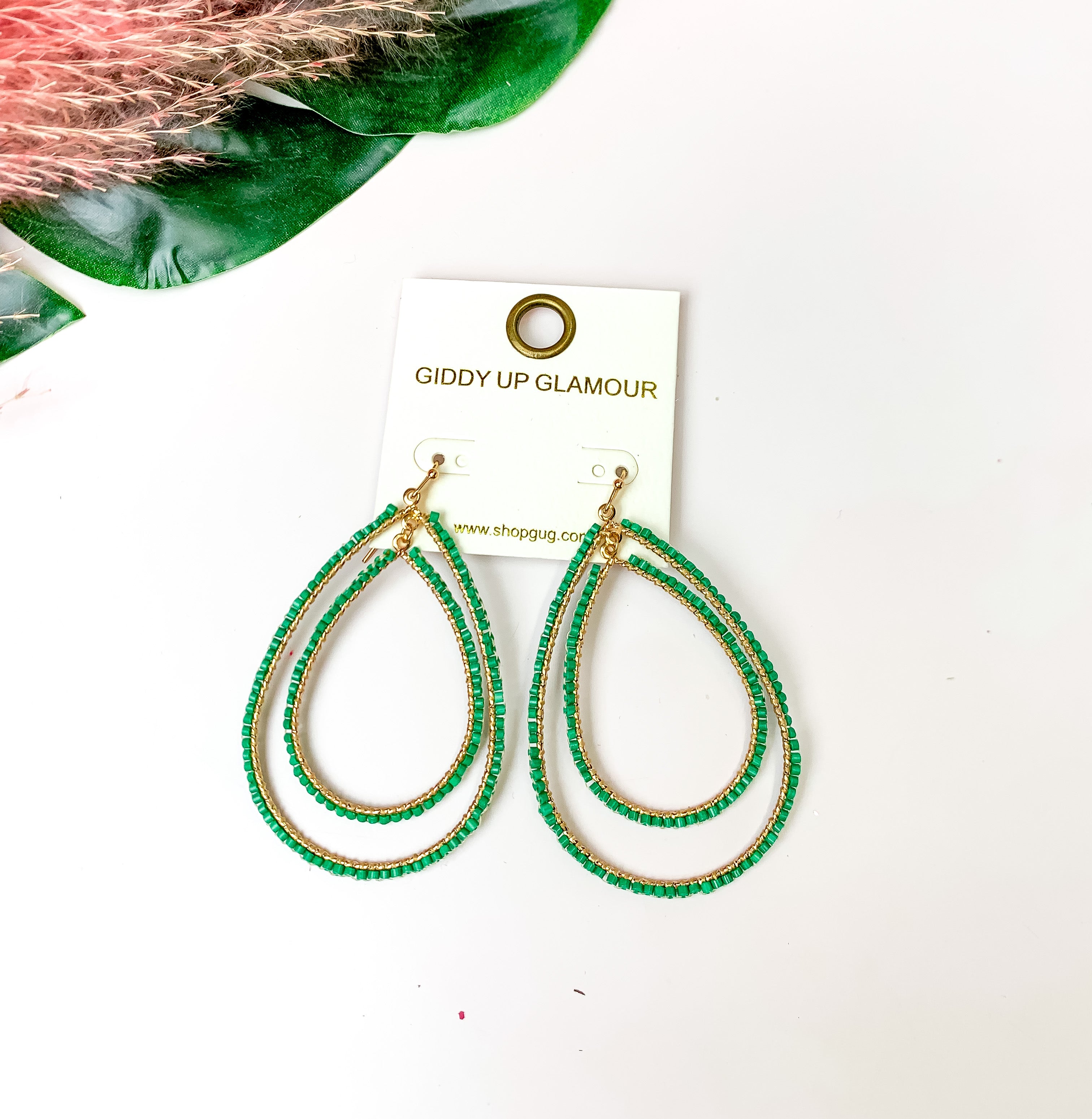 Double Open Teardrop Gold Tone Earrings with Beaded Outline in Green. Pictured on a white background with leaves in the top left corner.
