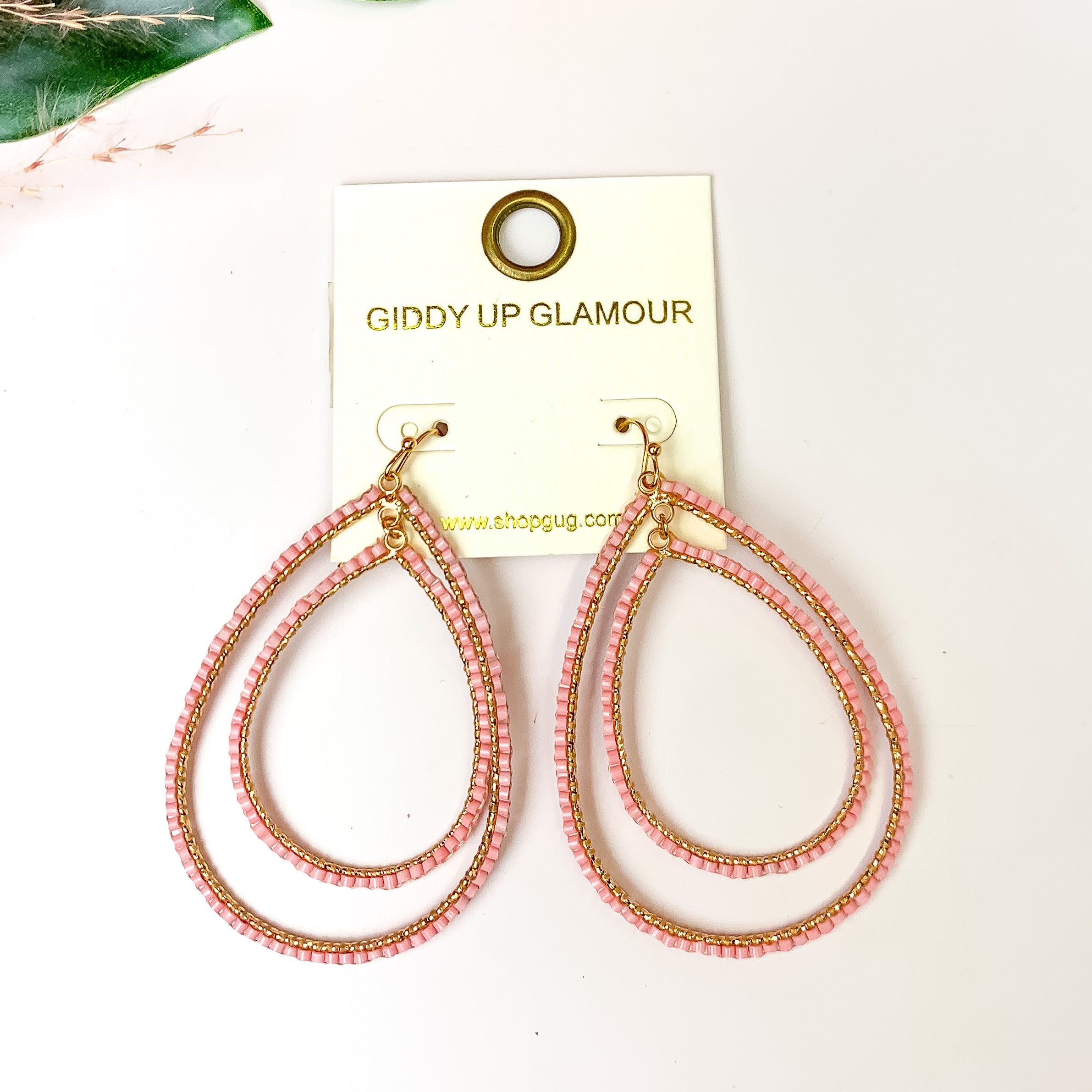 Double Open Teardrop Gold Tone Earrings with Beaded Outline in Light Pink. Pictured on a white background with leaves in the top left corner.