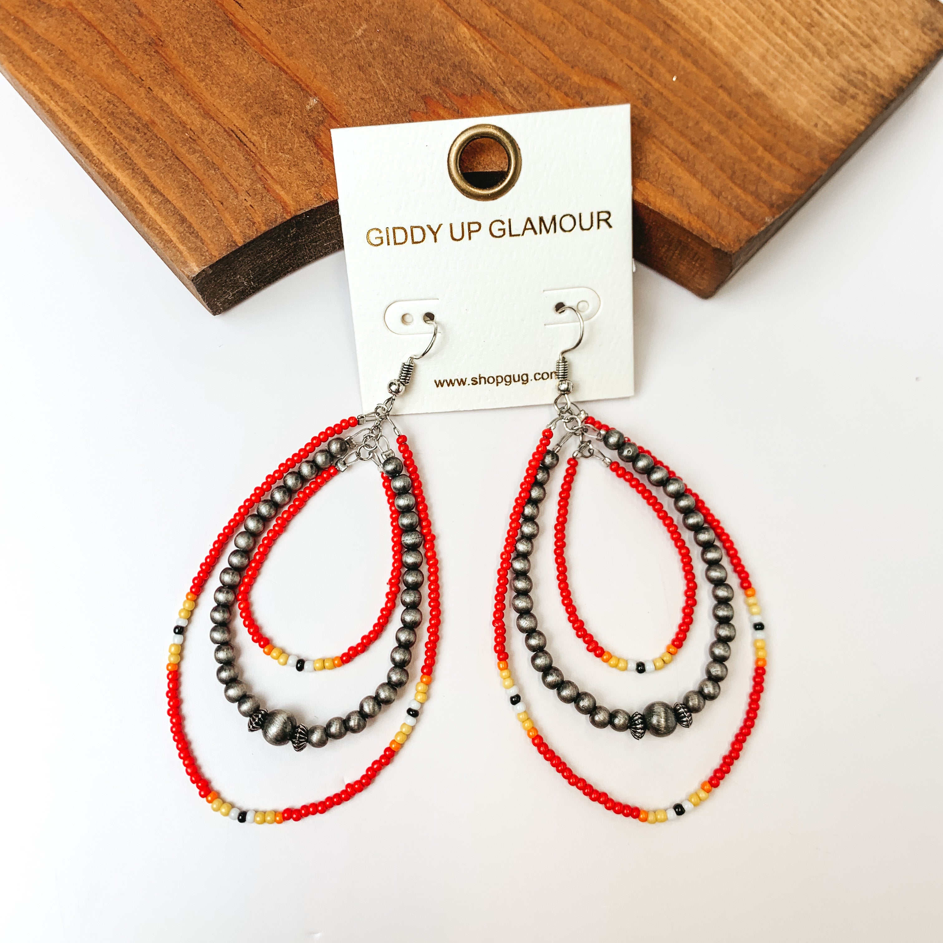 Western Style Triple Open Teardrop Earrings In Silver Tone and Red. Pictured on a white background with a wood piece behind the earrings.