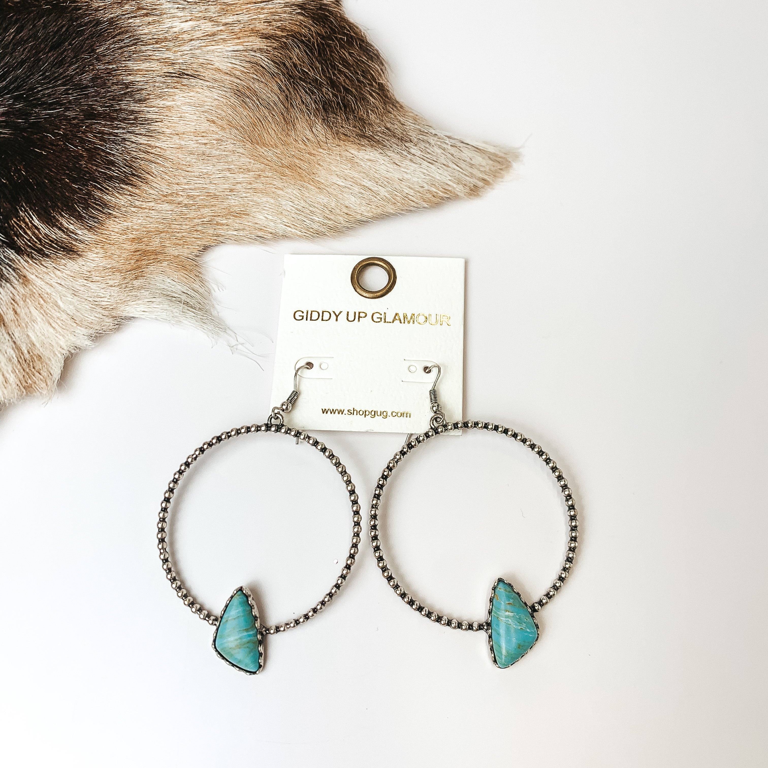 Silver Tone Textured Hoop Earring with Turquoise Stone. Pictured on a white background with faux animal skin on the top left corner. 