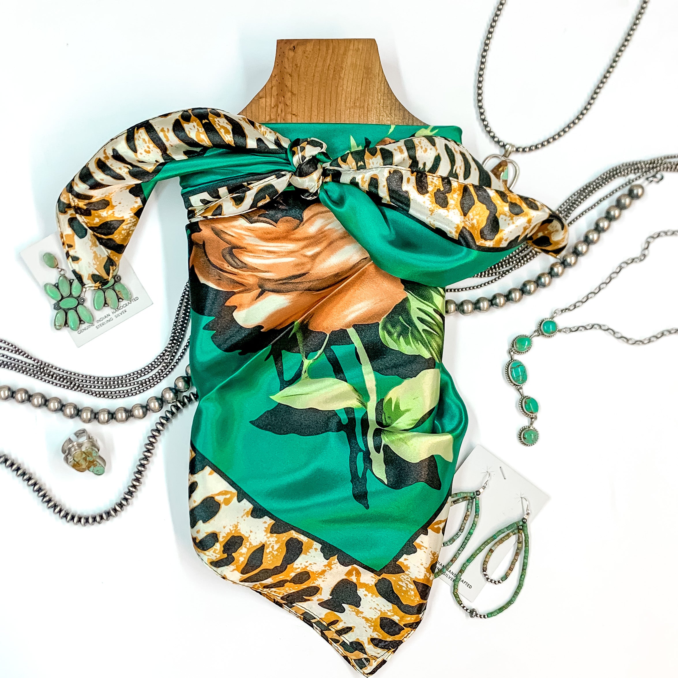 Patterned scarf in Fiesty Floral Emerald. Scarf is tied around a wooden piece. The scarf and piece of wood is pictured on a white background with Navajo jewelry spread out around it.