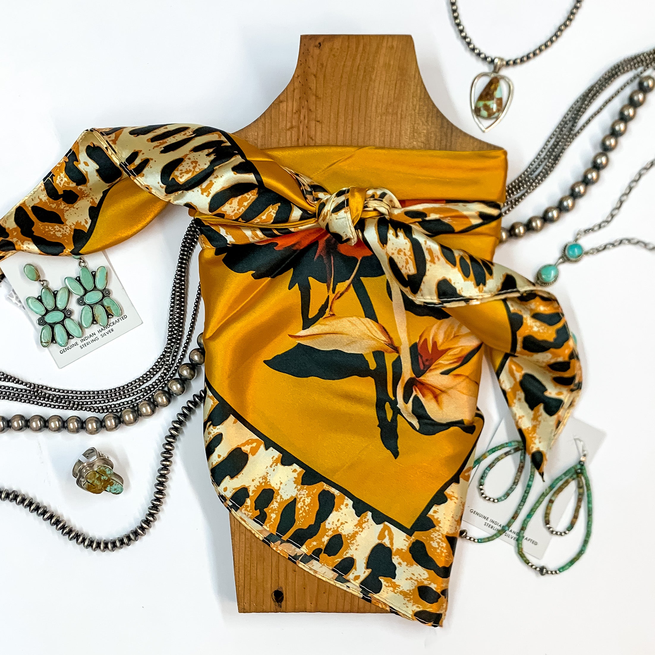 Patterned scarf in Fiesty Floral Mustard. Scarf is tied around a wooden piece. The scarf and piece of wood is pictured on a white background with Navajo jewelry spread out around it.